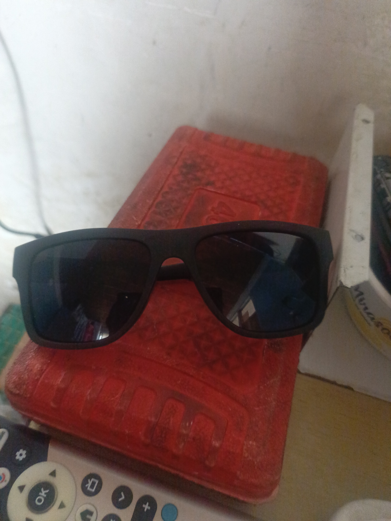 A pair of black sunglasses rests on a red, rectangular object. A remote control is partially visible in the foreground. The background is out of focus, but appears to be a white wall. I do not have information to determine if the image was taken in Juan Griego, Venezuela.