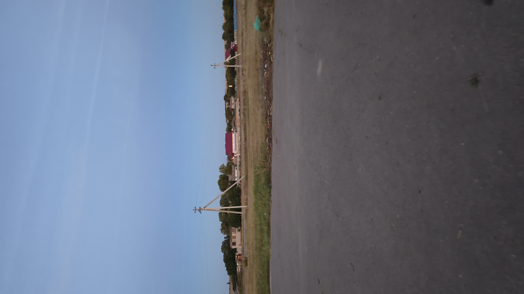 The image depicts a rural landscape under a clear blue sky. In the foreground, a paved road runs horizontally across the frame. Beyond the road, a grassy field with some scattered debris is visible. Further in the distance, a cluster of buildings, including houses with red roofs, are situated amidst trees and vegetation. Two utility poles with crossbars are prominent features in the middle ground. The scene suggests a quiet, possibly a village, setting.