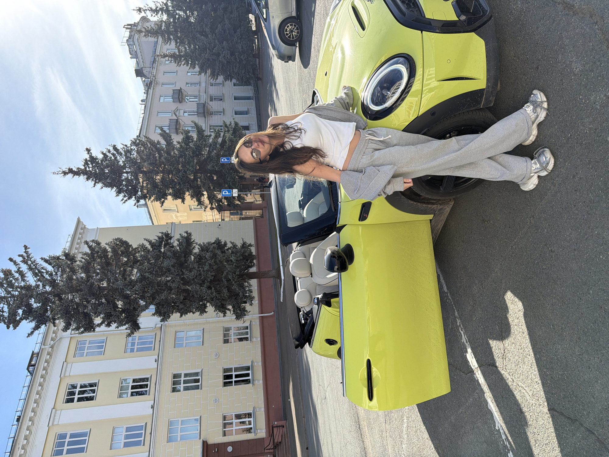 A woman with long brown hair and sunglasses is reclining on the hood of a yellow MINI Cooper convertible. She is wearing a white tank top and gray sweatpants, with a gray sweatshirt draped over her. The car is parked on a street near a building with multiple windows and trees, with a “P” parking sign visible. This location is Zheleznogorsk, Russia.