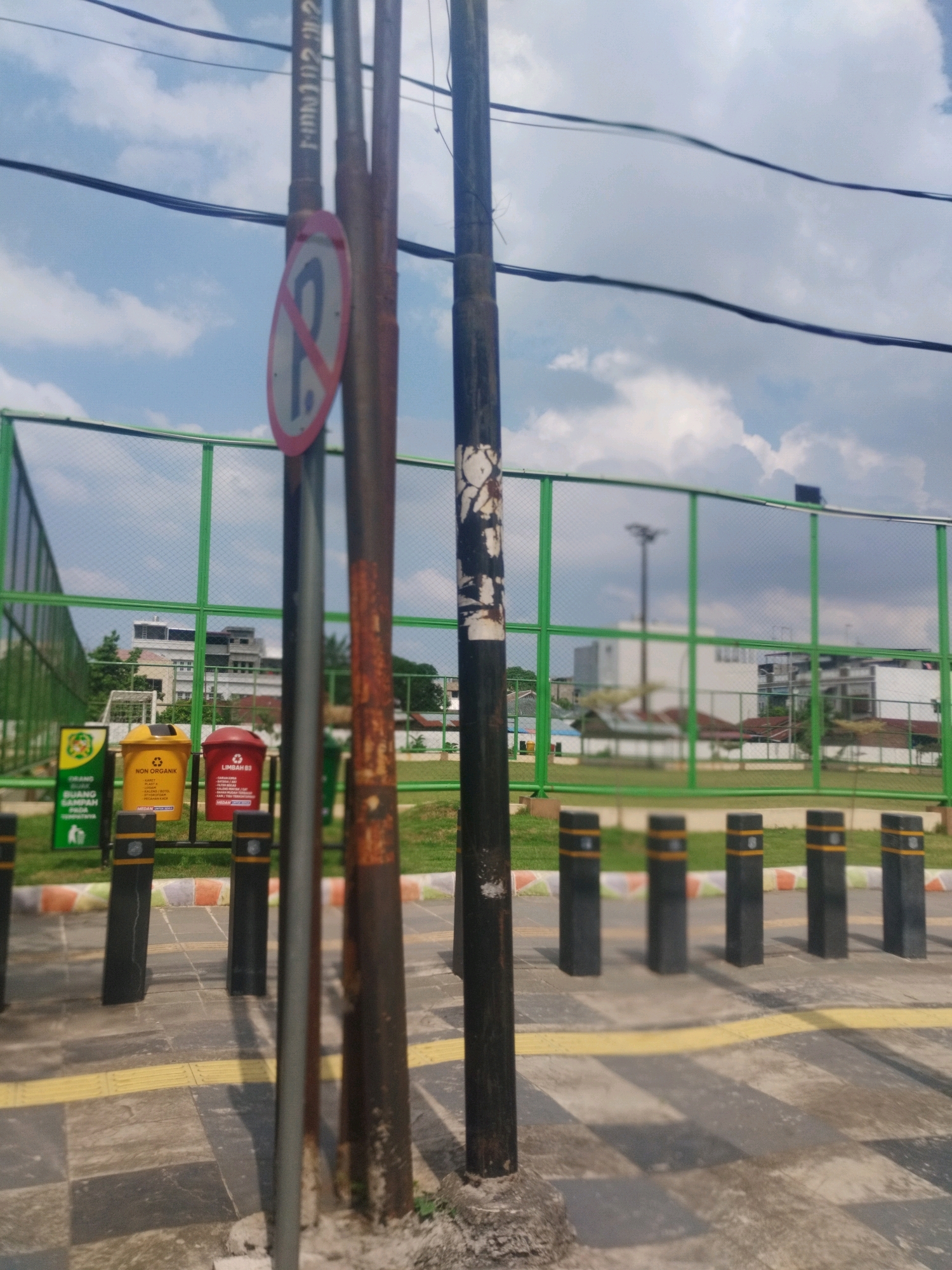 The image is an outdoor scene in the city of Medan, Indonesia, on a cloudy day. In the foreground, several metal poles stand vertically. One pole on the left displays a circular red and white sign with a diagonal line and the letter "P", indicating a no-parking zone. Next to this pole is a rusted metal pole. To the right, a black metal pole with peeling paint is visible. Behind these poles, a green mesh fence encloses an area with grass and trees. Beyond the fence, there are low-rise buildings and a larger, light-colored multi-story building. In front of the fence, several black bollards with yellow stripes are lined up along a paved walkway. Two colorful trash bins, one yellow and one red, are placed behind the poles and in front of the fence. The ground in the foreground is paved with interlocking blocks, with a yellow tactile paving strip running along the edge. The sky is filled with clouds.