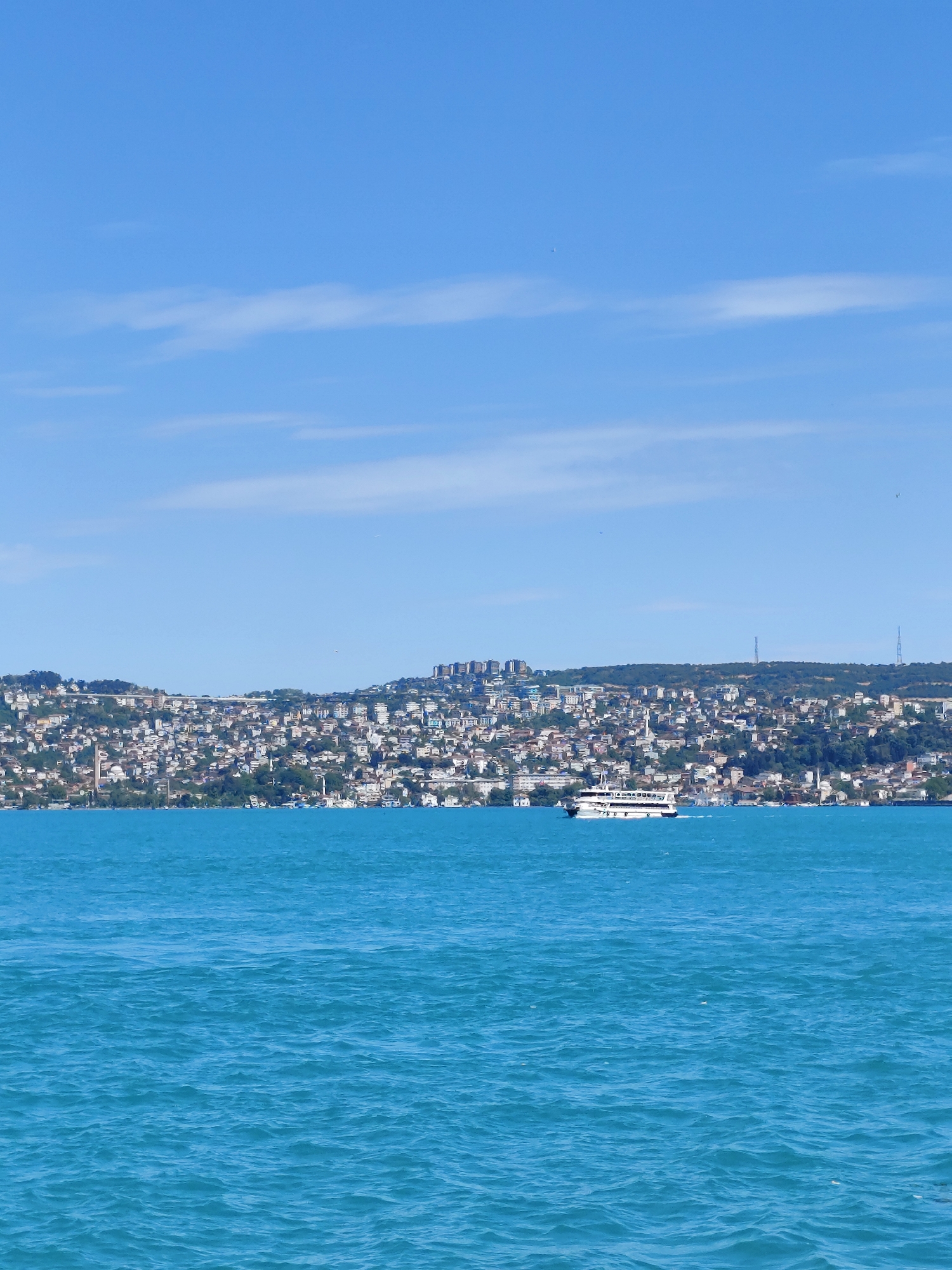 The scene is a daytime, medium-distance shot of the Sarıyer coastline in Istanbul, Turkey. The foreground is dominated by turquoise water with gentle ripples. In the middle ground, a densely populated hillside is visible, covered in closely packed buildings, mainly residential, interspersed with trees and vegetation. A white ferry moves along the coast. The sky above is light blue with scattered wisps of white clouds.