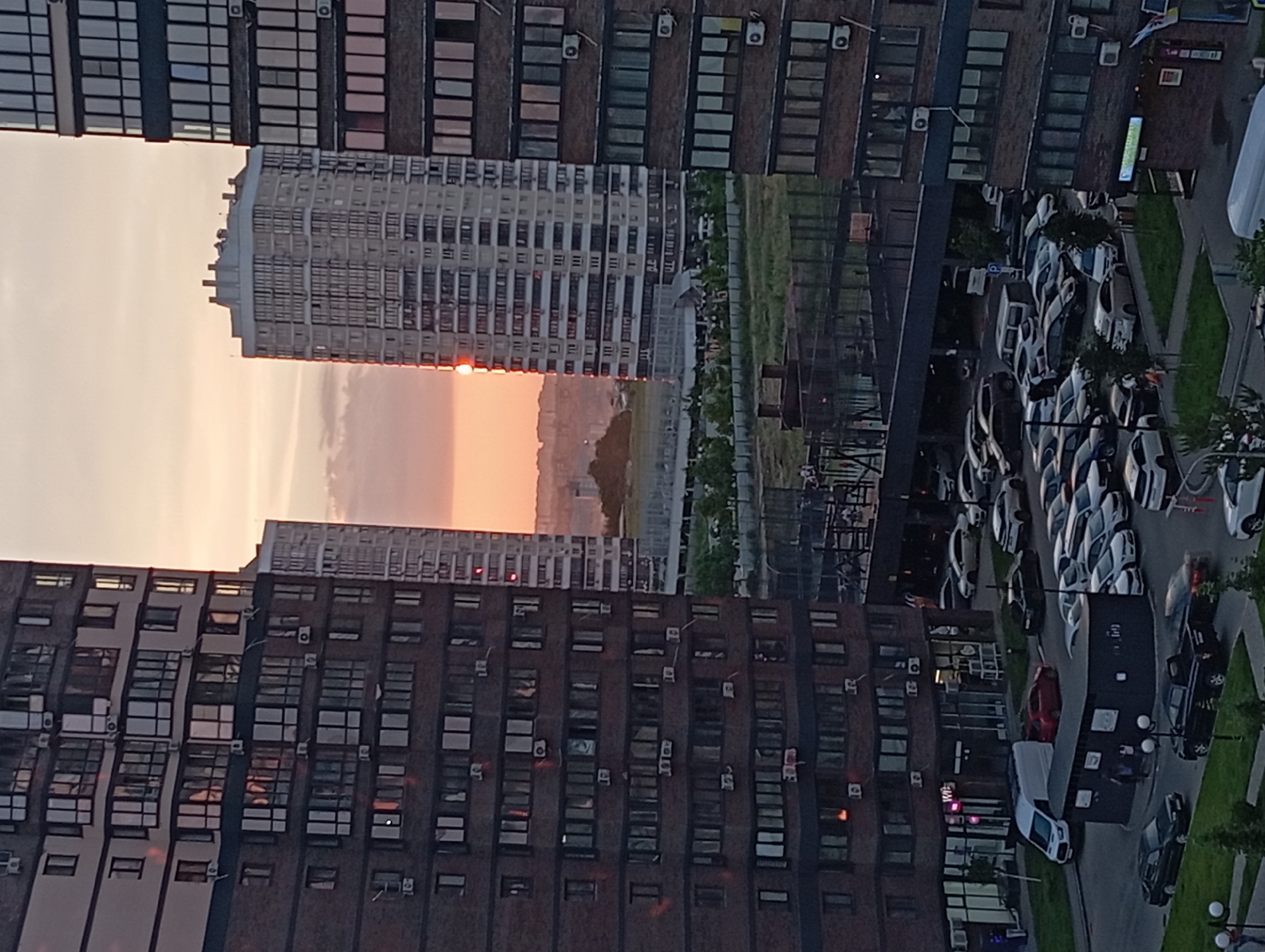 This image captures a sunset over a densely populated urban area in Krasnodar, Russia. In the foreground, a large, dark brick apartment building dominates the left side of the frame, its many windows reflecting the soft, warm light of the setting sun. To the right, another, lighter-colored apartment building stands, appearing taller and more modern. Between these structures, a third, very tall apartment building is visible in the distance, its facade a grid of windows. The sky is a gradient of orange, pink, and pale blue, with a few wispy clouds.
Below the buildings, a complex network of infrastructure is visible. A multi-lane road, lined with parked cars of various colors and models, winds through the scene. A white van is parked near the entrance of one of the buildings. In the mid-ground, a large, dark rectangular structure, possibly a commercial building or underground parking entrance, is situated, with a few people milling about its entrance. Beyond this, a fenced-off sports court or recreational area is discernible. Further into the distance, the hazy outlines of more buildings and infrastructure suggest a sprawling cityscape. The overall impression is one of a modern, active urban environment bathed in the peaceful glow of sunset.
