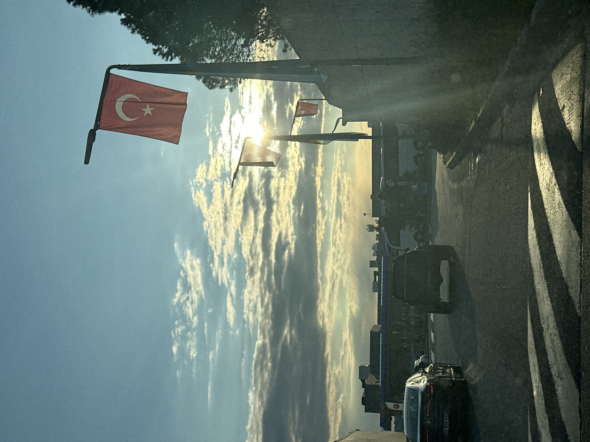The image shows a street scene in Gaziosmanpaşa, Turkey, with Turkish flags hanging from poles along the roadside. The sky is visible, with bright sunlight filtering through the clouds. Several cars are on the street, including a dark-colored car in the foreground. The right side of the frame shows a sidewalk with pedestrian markings. Buildings and other infrastructure are visible in the background.