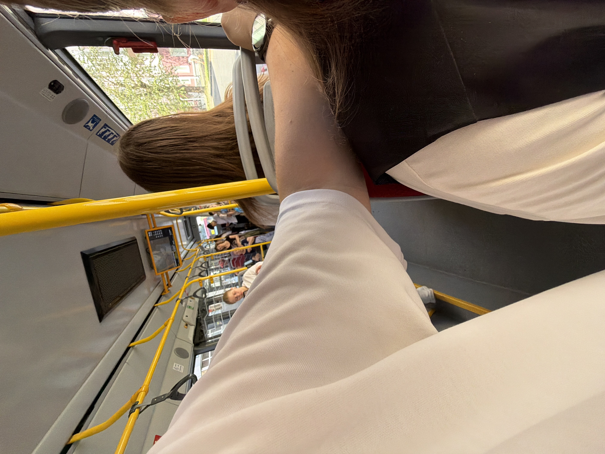 The image shows the interior of a bus, likely in Perm, Russia, based on context clues. In the foreground, a person wearing a white polo shirt is visible, with their arm extended across the frame. To their right, another person is seated, wearing a black vest over a white shirt. The bus has yellow grab poles and handrails throughout the cabin. Passengers are seated and standing in the background, with one person in a white top and another in a black top and striped skirt visible. The window shows a street scene with trees and buildings, suggesting the bus is in motion or stopped at a light. The overall environment is typical of public transportation.