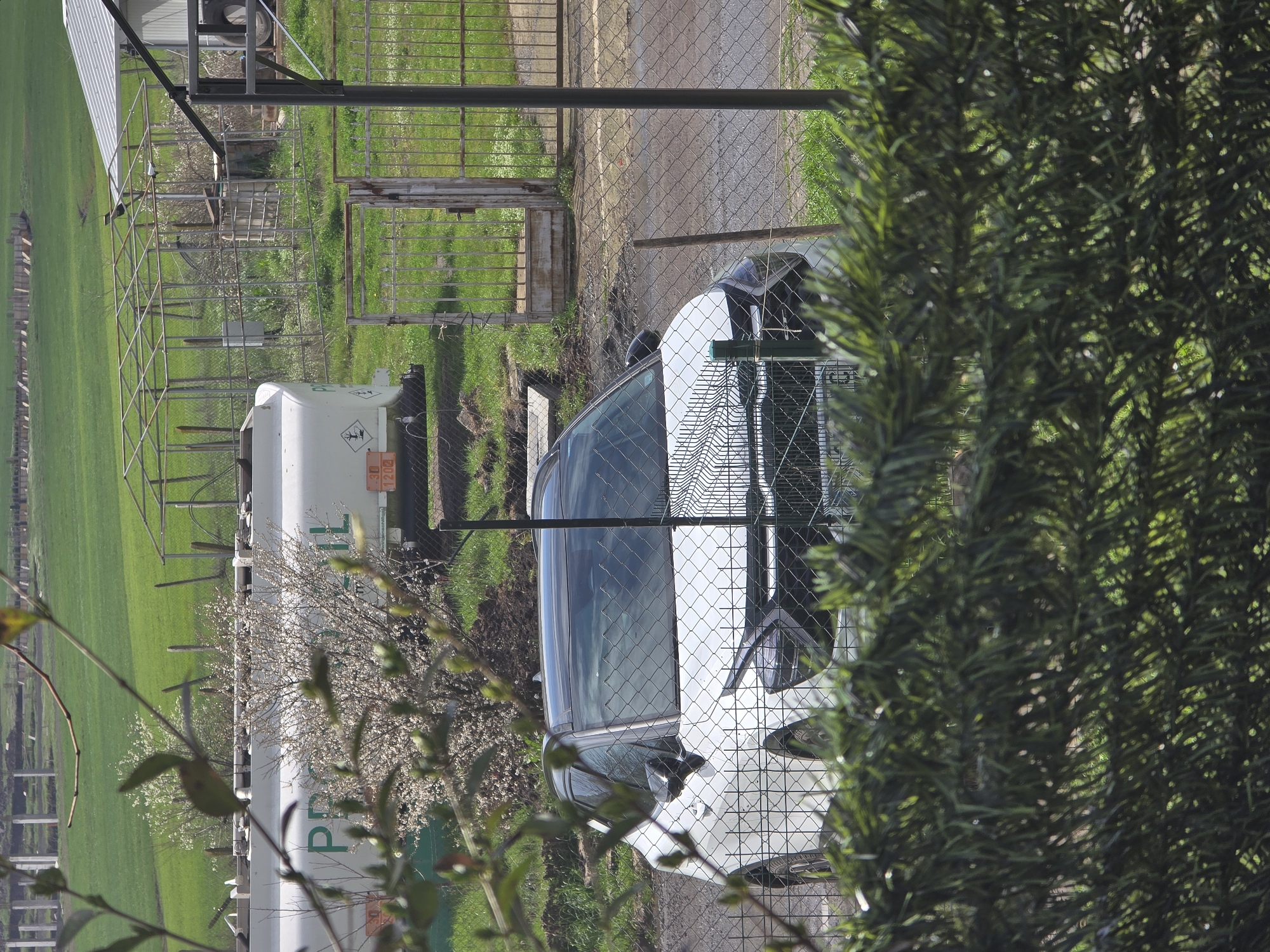 The image shows a white car parked near a fence and gate, with green vegetation and a tank in the background. The scene appears to be in a rural or semi-rural area, and the location in Burgas, Bulgaria is not apparent from the image.