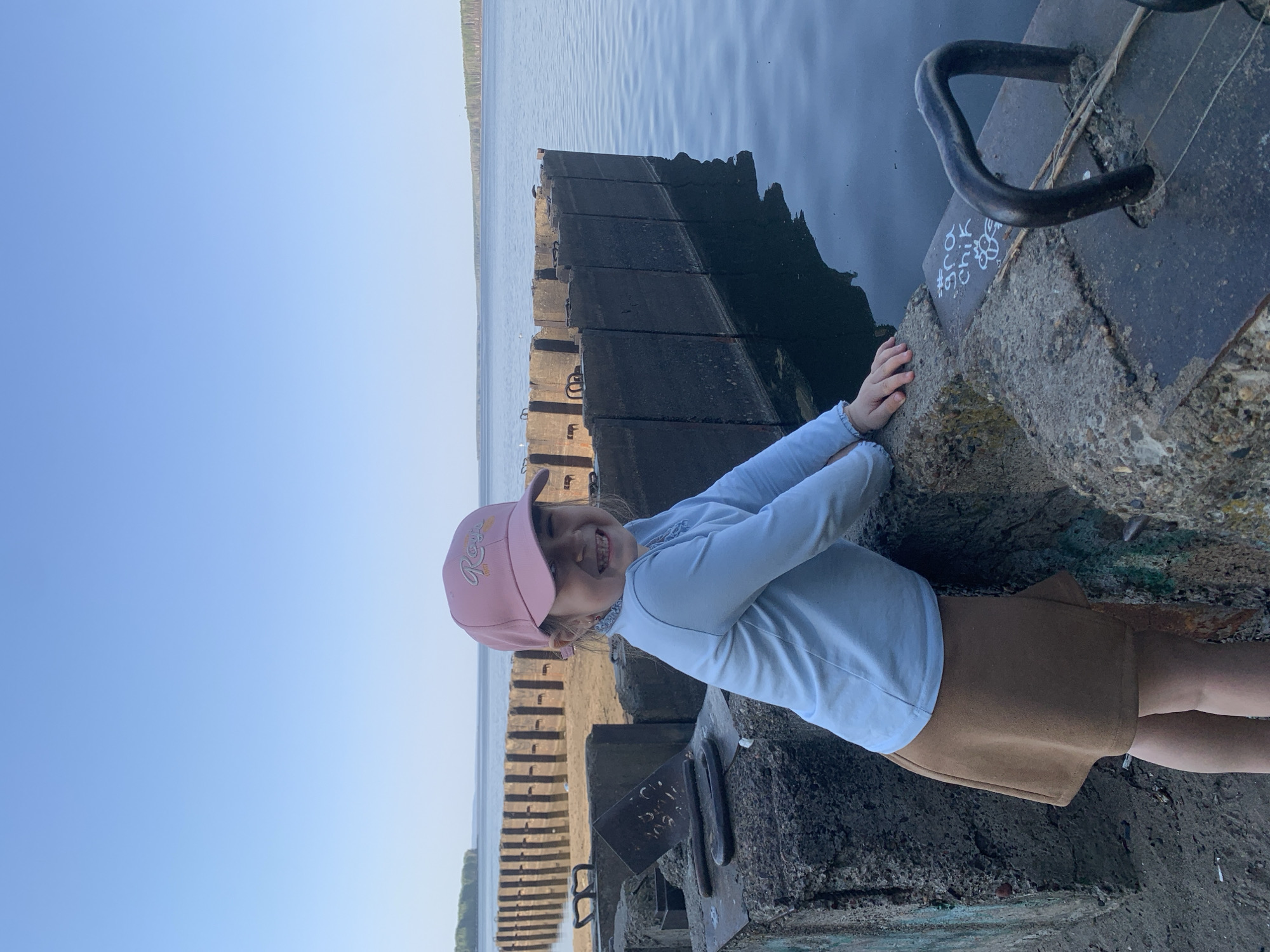 A young girl stands on a concrete pier, with a backdrop of water and the sky. She is wearing a pink baseball cap, a light blue long-sleeved shirt, and a brown skirt. Her hands are resting on the edge of the pier. The pier is made of rough, weathered concrete and includes metal hardware. Beyond the pier is a body of water, likely a river or lake, and in the distance, there appears to be a tree line. The sky is clear and blue. The photo was taken in the city of Chaikovsky, Russia.