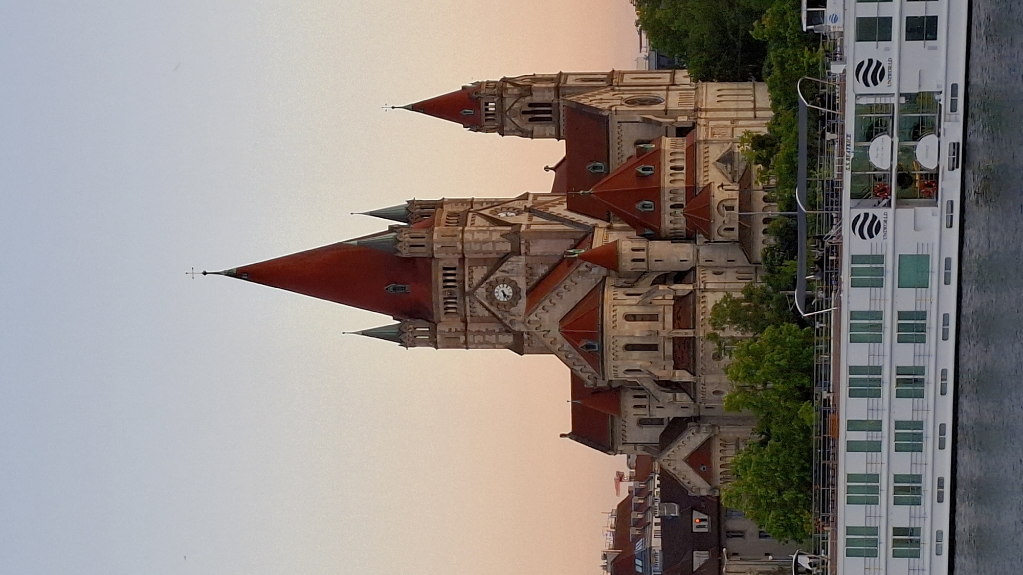 The image depicts the Votivkirche in Vienna, Austria, situated next to a river or canal. The church is a large, ornate structure with a reddish-brown roof and multiple spires. A clock is visible on one of the towers. Adjacent to the church, partially obscured by trees, is a large white river cruise ship with numerous windows. The sky is a gradient of light blue and pink, suggesting either sunrise or sunset. The image appears to have been taken from a slightly lower vantage point, possibly from another vessel on the water.
