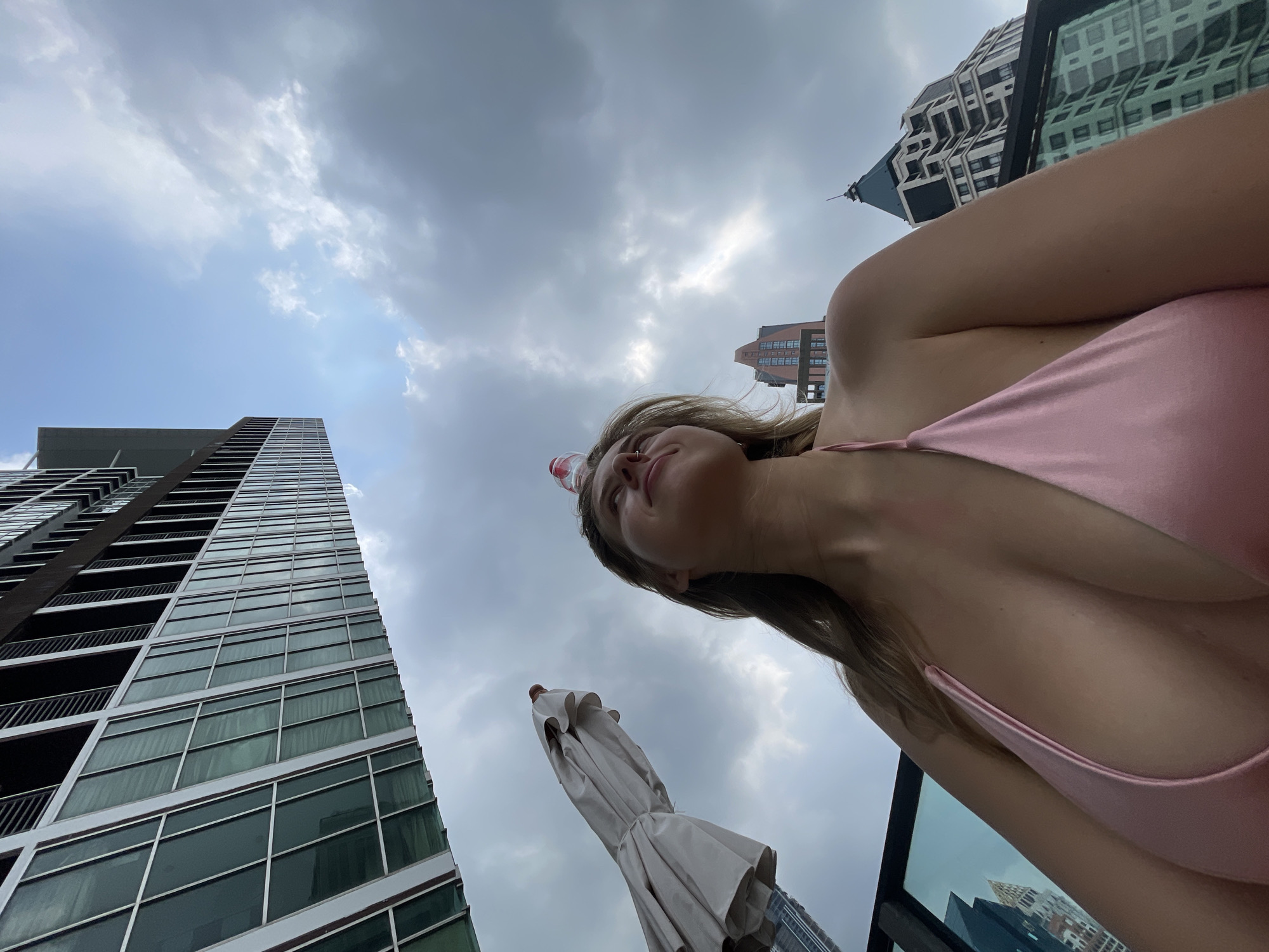 The image captures a worm's-eye view of a woman and the surrounding city skyline under a cloudy sky. The primary subject, a woman with blonde hair, is positioned in the lower right quadrant, looking upwards with a serene expression. She is wearing a pink top or swimsuit, and a small, clear plastic water bottle with a red cap is balanced playfully on her forehead.
The scene is set in an urban environment, likely from a high-rise balcony or rooftop, offering expansive views of modern architecture. To the left, a tall, glass-faced skyscraper with numerous balconies extends vertically into the frame. In the middle-ground, a closed beige patio umbrella stands, also viewed from below. In the distance, several other distinct buildings with contemporary designs are visible against the backdrop of an overcast sky with patches of brighter light breaking through the clouds. The architecture is consistent with a bustling modern metropolis, suggesting a location like Bangkok, Thailand.
The time of day is daytime, indicated by the overall brightness despite the cloud cover. The weather is cloudy but not stormy. No activity beyond the woman gazing upwards is explicitly depicted, conveying a sense of relaxation or contemplation. There is no visible text in the image.
