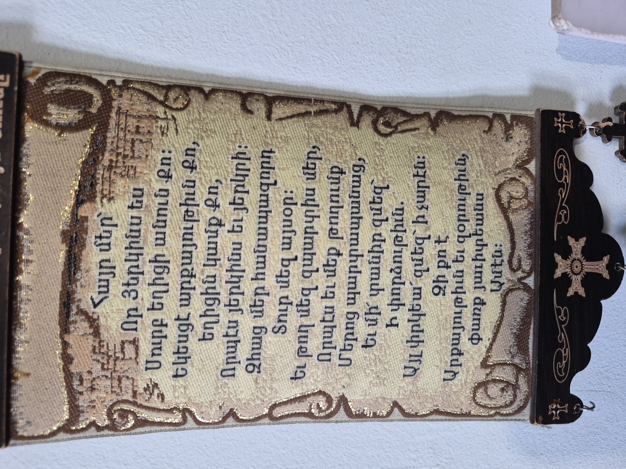 The image displays a decorative textile wall hanging or banner, oriented vertically against a plain white background, likely an indoor wall. The banner features a cream or beige woven fabric adorned with intricate embroidery in brown and shimmering gold threads.
The central part of the banner contains the Lord's Prayer (Hayr Mer) in Armenian script, embroidered in brown. The text is framed within an elaborate, scroll-like design, also outlined with golden thread. The borders of the banner are decorated with ornate, swirling patterns in gold and brown.
Visible text:
"Հայր մեր՝
Որ Յերկինս ես
Սուրբ եղիցի անուն Քո։
Եկեսցէ արքայութիւն Քո,
եղիցին կամք Քո,
Որպէս յերկինս եւ յերկրի։
Զհաց մեր հանապազորդ
Տուր մեզ այսօր։
Եւ թող մեզ զպարտիս մեր,
Որպէս եւ մեք թողումք
Մերոց պարտապանաց,
Եւ մի՛ տանիր զմեզ`
Ի փորձութիւն.
Այլ փրկեա՛ զմեզ` ի չարէն։
Զի քո է
Արքայութիւն եւ զօրութիւն,
փառք յաւիտեան
Ամէն։"
To the right edge of the banner, a dark, possibly wooden or metal, decorative element with two stylized Armenian crosses is attached, suggesting it might be part of the hanging's frame or a separate religious accessory. The crosses feature lighter, possibly golden, inlays or etched designs. Small metal hooks or rings connect this darker piece to the embroidered banner. The textile exhibits a visible weave pattern, indicating a woven fabric base with machine or hand embroidery. No people or specific activity are depicted; the image focuses solely on this religious and decorative artifact.