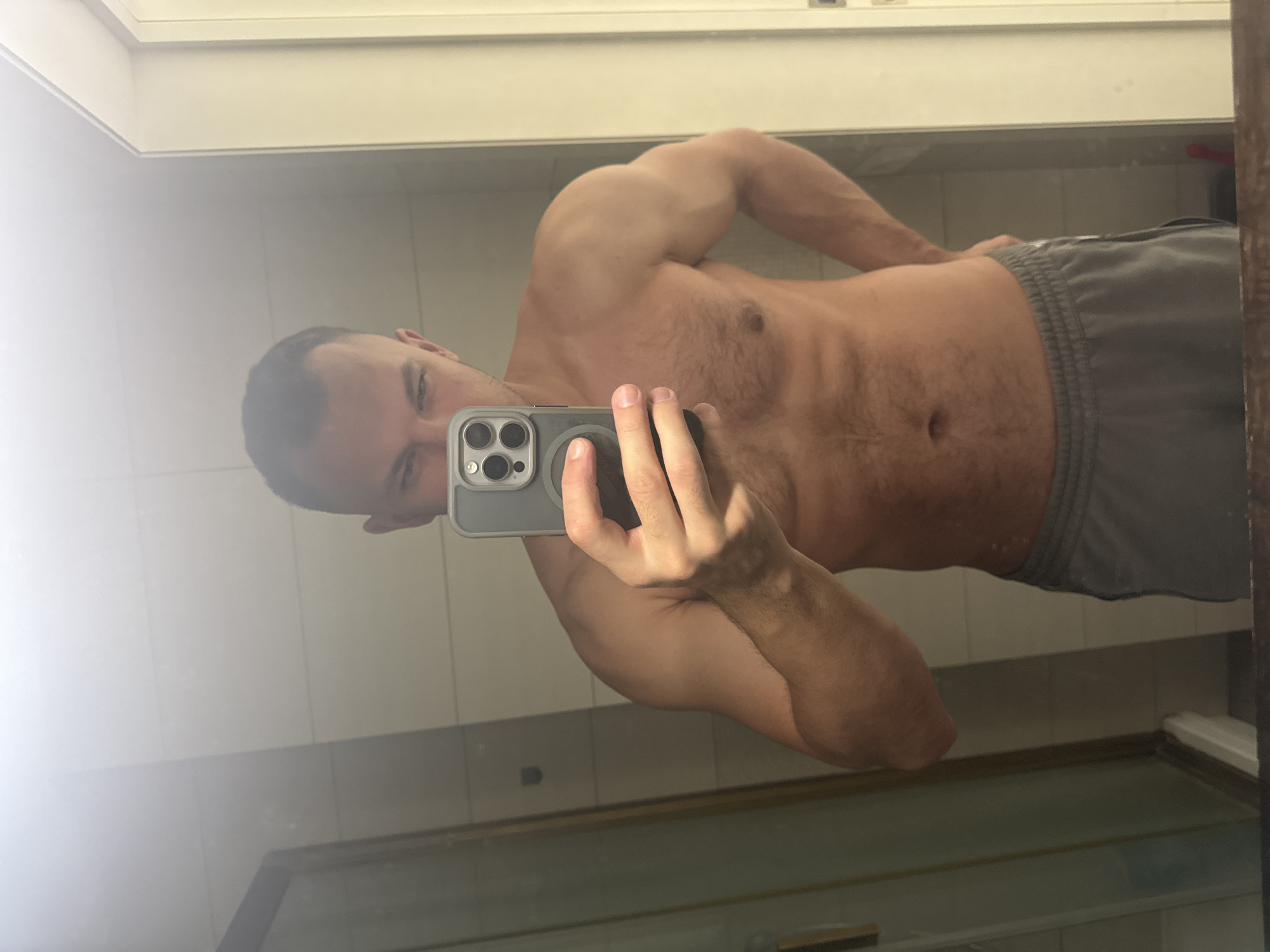 A shirtless man is taking a selfie in a bathroom mirror. He is wearing gray athletic shorts. The location is not discernible from the image.