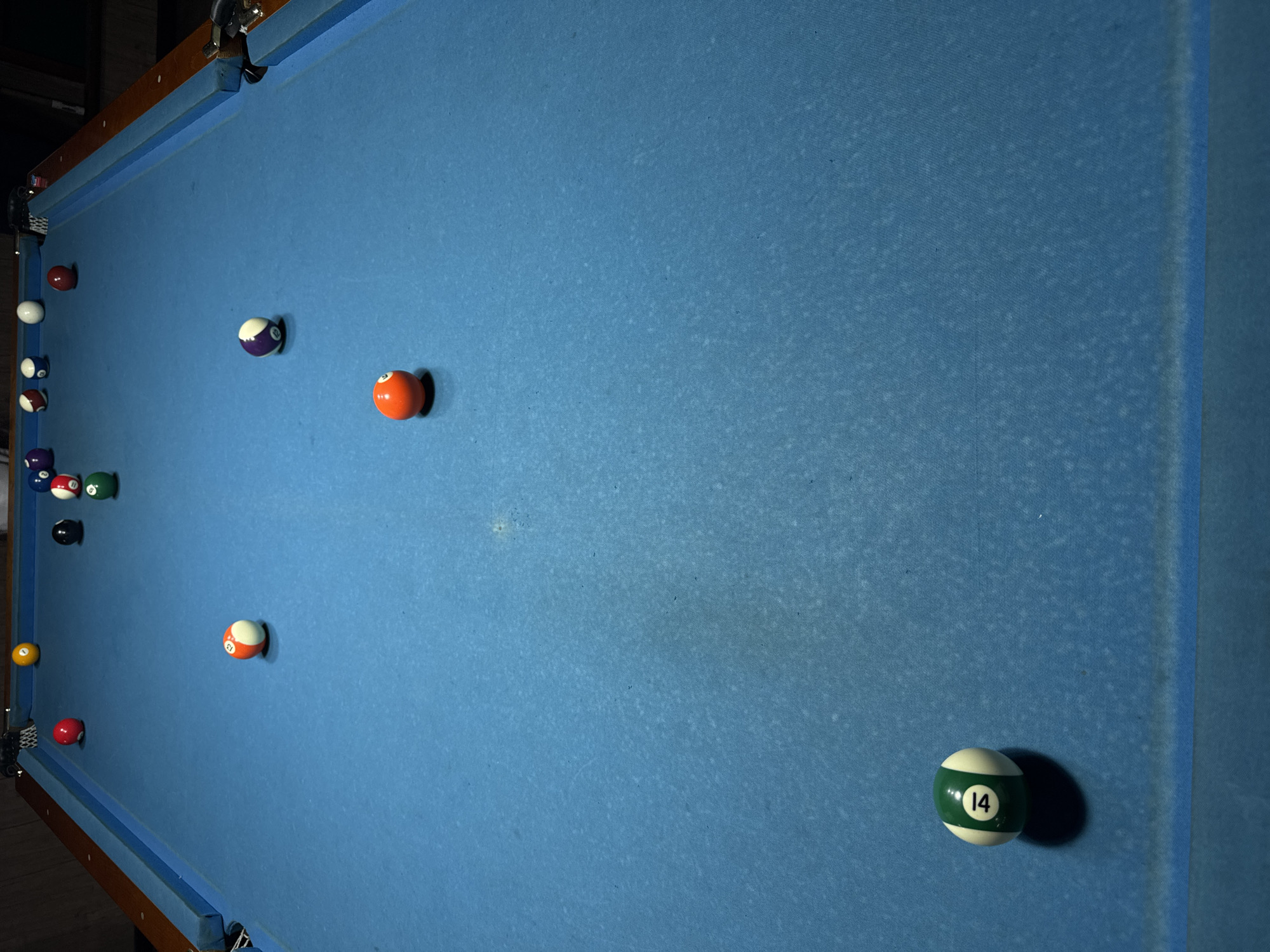 This is a medium-shot, eye-level, wide-angle photograph of a blue billiard table. The table is set up with a collection of billiard balls, suggesting a game is either in progress or has recently concluded. The lighting suggests it is daytime, possibly indoors in a dimly lit room or entertainment venue.
Several billiard balls are scattered across the table. Visible balls include a solid orange ball with no number, a striped ball with orange and white sections and the number "13", a striped ball with purple and white sections, a solid red ball with no number, a solid green ball with no number, a solid yellow ball with no number, a striped ball with green and white sections and the number "14", and a solid dark blue ball with no number. There are also other balls partially obscured or less distinct in the image. The blue felt of the table is textured and shows some signs of wear, with scattered light-colored spots and dust. The wooden edge of the billiard table is visible along the left and top edges of the frame. The background on the far left appears to be a dark, possibly wooden, wall or furniture.
The scene is static, with no people visible. The arrangement of the balls suggests a game of pool is being played, or has been played. The setting appears to be an indoor recreational space. No specific time of day or weather conditions are discernible beyond the artificial lighting. No text is clearly visible other than the numbers on the billiard balls themselves ("13" and "14"). The location context of Tashkent, Uzbekistan, does not provide specific visual cues within the image itself.