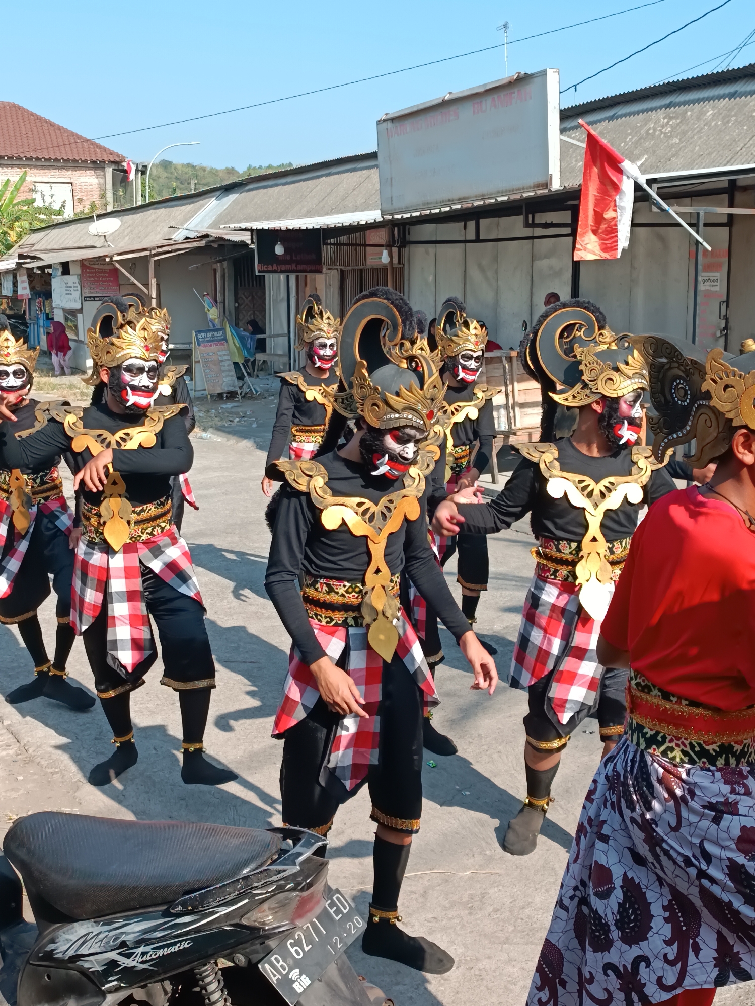 A group of performers in traditional Balinese dance costumes are walking down a street in Imogiri, Indonesia. The performers are wearing black long-sleeved shirts, black pants, and red and white checkered sarongs. They also have on elaborate gold headdresses and masks with white and red face paint. The street is paved and lined with buildings, some of which have corrugated metal roofs. A red and white Indonesian flag is visible on a pole in the background. A motorcycle is parked in the foreground on the left side of the image. The sky is clear and blue.