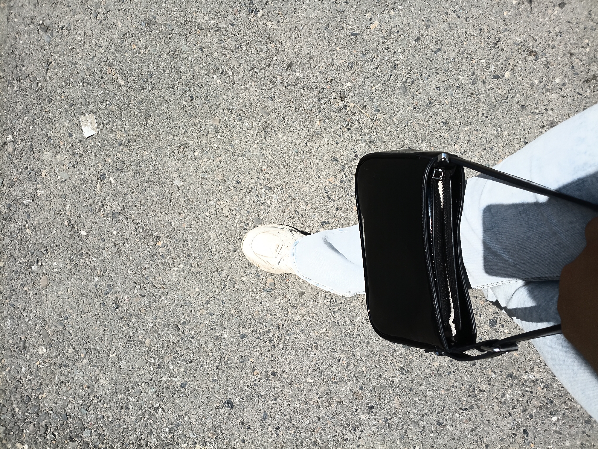 The image shows a person's leg, foot, and a black handbag on an asphalt surface. The person is wearing light blue jeans and white sneakers. The handbag is glossy black with a zipper closure and a strap. The asphalt is gray and speckled with small stones. A small white piece of litter is visible on the asphalt to the left of the foot. The lighting suggests it is daytime, with shadows indicating sunlight. The scene appears to be an outdoor urban environment, possibly a street or sidewalk in Osh, Kyrgyzstan.