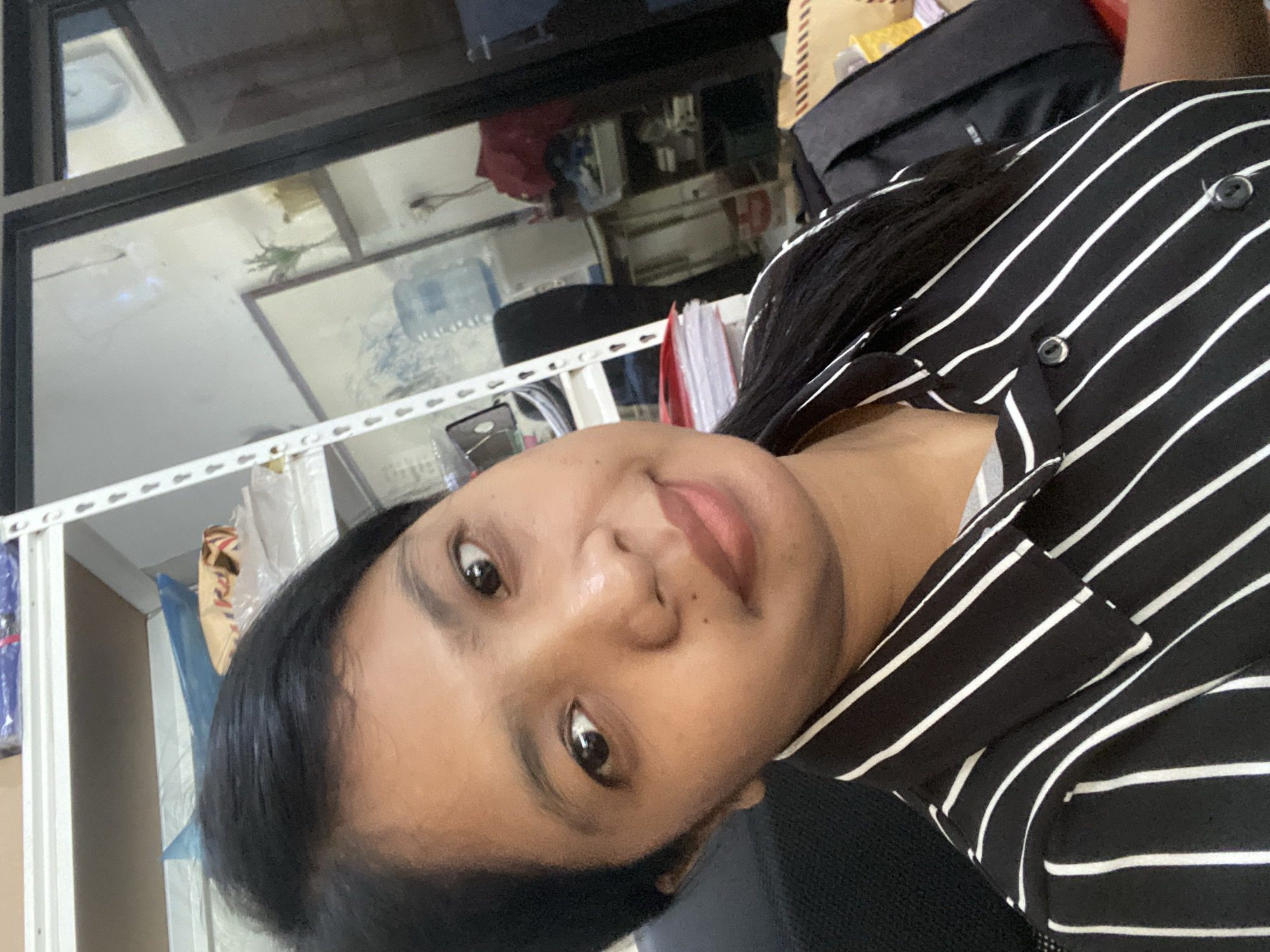 A medium-close-up indoor shot features a woman with dark hair wearing a black and white striped shirt. The background includes shelves holding various objects, office supplies, and a window. A dark bag is also partially visible. The location is potentially in Negara, Indonesia.