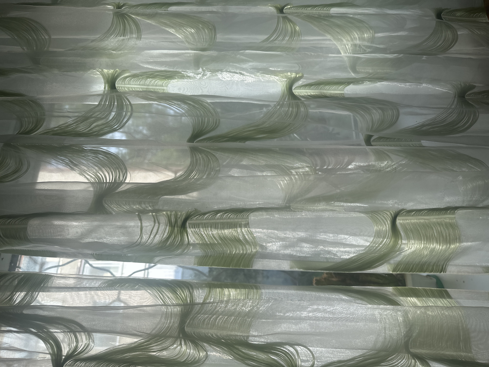 This media file features a close-up view of a white sheer fabric with a subtle green textured pattern, resembling strands of hair or flowing ribbons. The fabric is layered, creating horizontal bands of light and shadow. The pattern appears to be embroidered or woven into the sheer material, with each section of the pattern consisting of numerous fine, parallel strands that curve and interlace.
The setting appears to be indoors, with natural light filtering through the fabric, suggesting it's a window covering or decorative textile. Through the sheer material, faint outlines of what looks like a window frame and possibly the outdoors can be discerned, hinting at a domestic or office environment. The time of day is indeterminate but suggests daylight hours due to the quality of light. No people, specific activities, or emotions are discernible. No visible text is present in the image. The location context of Togliatti, Russia, does not offer any specific visual cues within the image itself. The overall impression is one of delicate texture and diffused light.