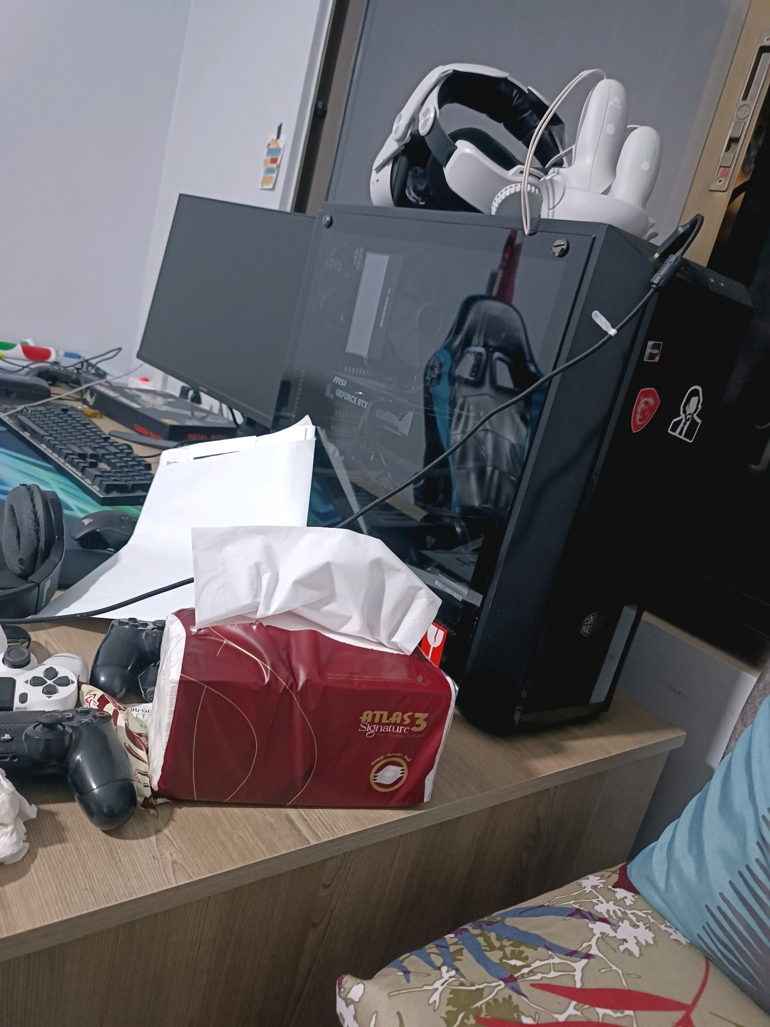 The image depicts a cluttered desk in a room, likely in the city of السعادة, Jordan. On the desk, a computer tower with a glass side panel is visible, showcasing internal components and a black gaming chair reflected inside. To the left of the computer, a dark monitor is present. Various gaming accessories are scattered around, including two game controllers (one white and one black), headphones, and a mouse. A box of tissues with a red and gold wrapper sits prominently in the foreground. In the background, a white VR headset and controllers are placed on top of the computer tower. The desk itself is made of a light-colored wood. The overall impression is of a gaming or computer enthusiast's workspace, characterized by disarray and a focus on technology and gaming.