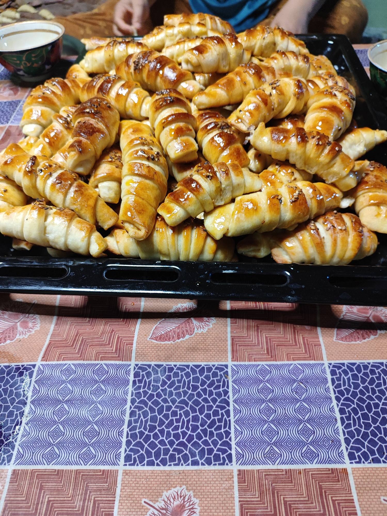 A close-up, top-down shot shows a baking tray filled with golden-brown crescent-shaped pastries, likely croissants or rugelach, sprinkled with sesame seeds. The pastries are arranged in multiple layers on a black baking sheet, which rests on a table covered with a patterned cloth featuring geometric designs in shades of purple, pink, and beige. In the upper left corner, a partially visible decorative cup containing a light-colored liquid is present. The background is slightly blurred, showing a hint of fabric and a person's hand. The overall scene suggests a domestic setting where baked goods are being prepared or served, possibly in Ismoili Somoni, Tajikistan, given the distinctive patterns on the tablecloth.