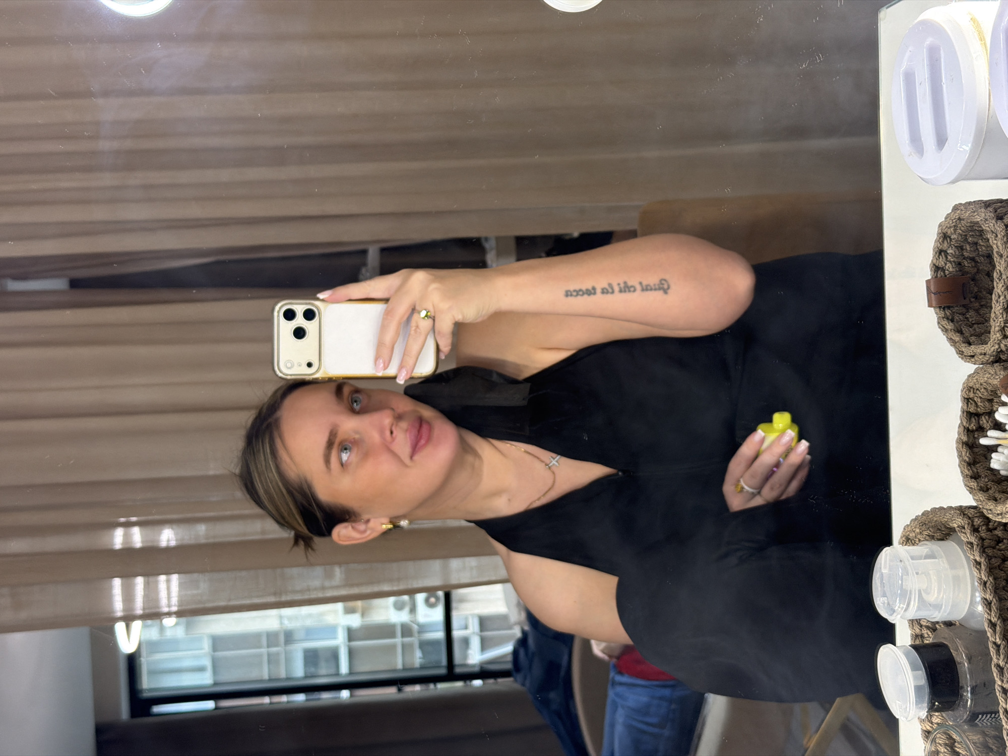 A woman is captured in a mirror selfie, holding a light-colored smartphone with a prominent camera array in her left hand (her right hand, reflected) to take the photo. She has dark hair styled back, light-colored eyes, and wears a black top or dress with a V-neckline, accessorized with a subtle cross necklace and a small pearl earring. On her left arm (her right arm, reflected), a tattoo is visible. In her right hand (her left hand, reflected), she holds a small, yellow and white object, possibly a vape pen or a cosmetic product.
The background reflected in the mirror features light brown vertical blinds or wooden panels, behind which a window reveals urban buildings, suggesting an indoor city setting during the daytime. Part of another person in a blue shirt is faintly visible in the lower right background reflection. To the right side of the mirror, a white shelf holds various small, clear plastic bottles with white or black caps, and a brown crocheted basket containing what appears to be cotton swabs.
The woman's expression is casual and relaxed, with a slight smile as she looks towards the phone/mirror. Her fingernails have a French manicure style, and she wears a ring on her ring finger.
**Visible text:** The tattoo on her left arm (her right arm, reflected) reads "dlu ad al tacol" in mirrored text.