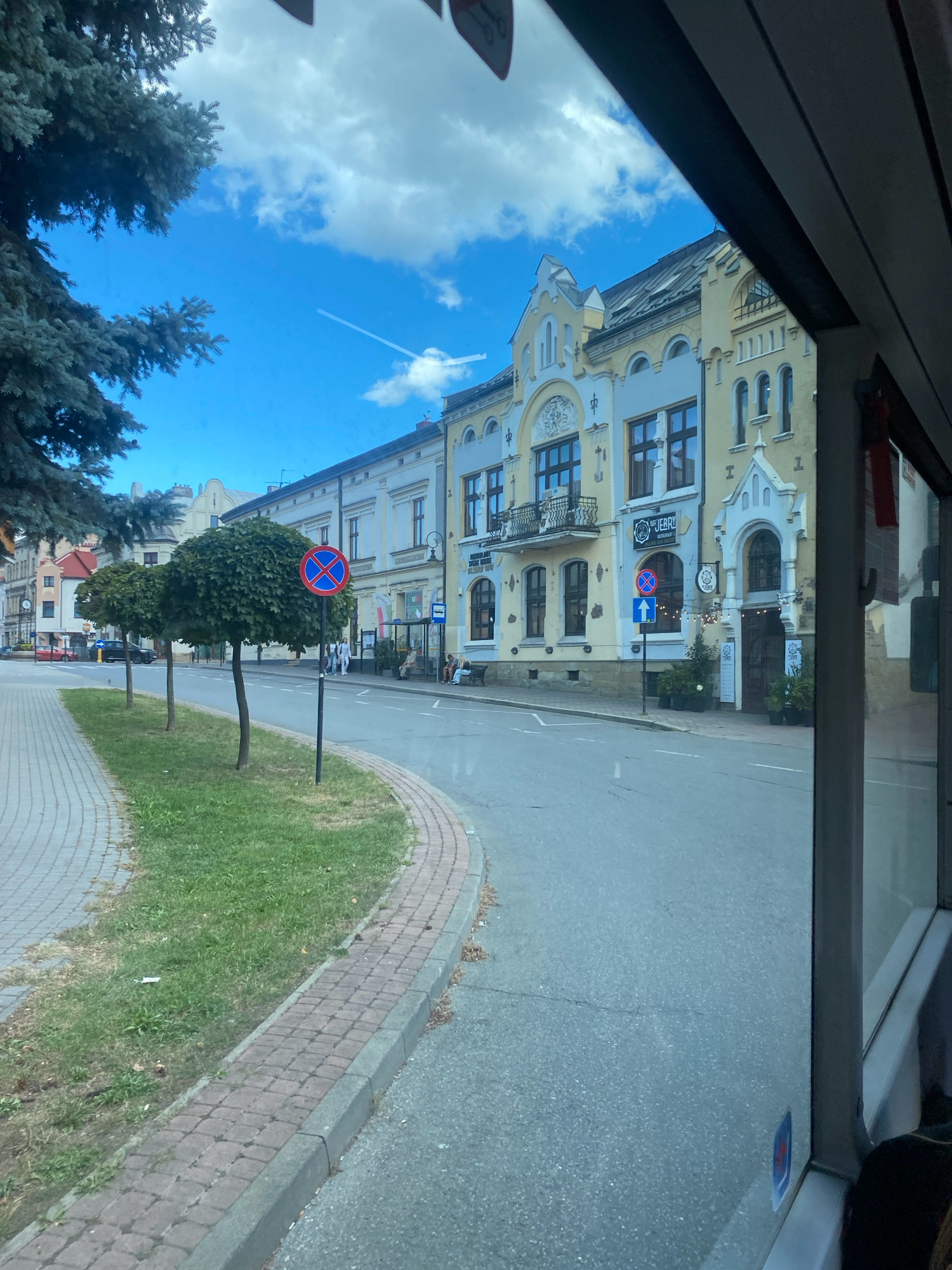 The image captures a street scene in Nowy Sącz, Poland, viewed from the interior of a vehicle, likely a bus, given the perspective and the presence of a window frame. The street is paved and curves gently to the left. To the left of the street is a sidewalk made of brick pavers, bordered by a concrete curb and a strip of green grass. Several trees with rounded canopies are planted in the grassy area along the sidewalk.
Across the street, a row of buildings with light-colored facades, predominantly yellow and white, lines the road. These buildings exhibit architectural details typical of European cities, with multiple stories, arched windows, and decorative elements. Some of the buildings appear to house businesses, with signs visible, including one that reads "MUJERRI RESTAURANT".
Several traffic signs are present. One is a circular red and blue sign indicating "no parking" or "no stopping". Another sign below it shows a blue arrow pointing upwards. Further down the street, a bus stop shelter is visible, with people seated on a bench.
The sky is blue with scattered white clouds, and a contrail from an aircraft is visible. A large evergreen tree is situated on the far left of the frame, partially obscuring the sky and some of the buildings. The overall impression is of a pleasant day in a European urban environment.