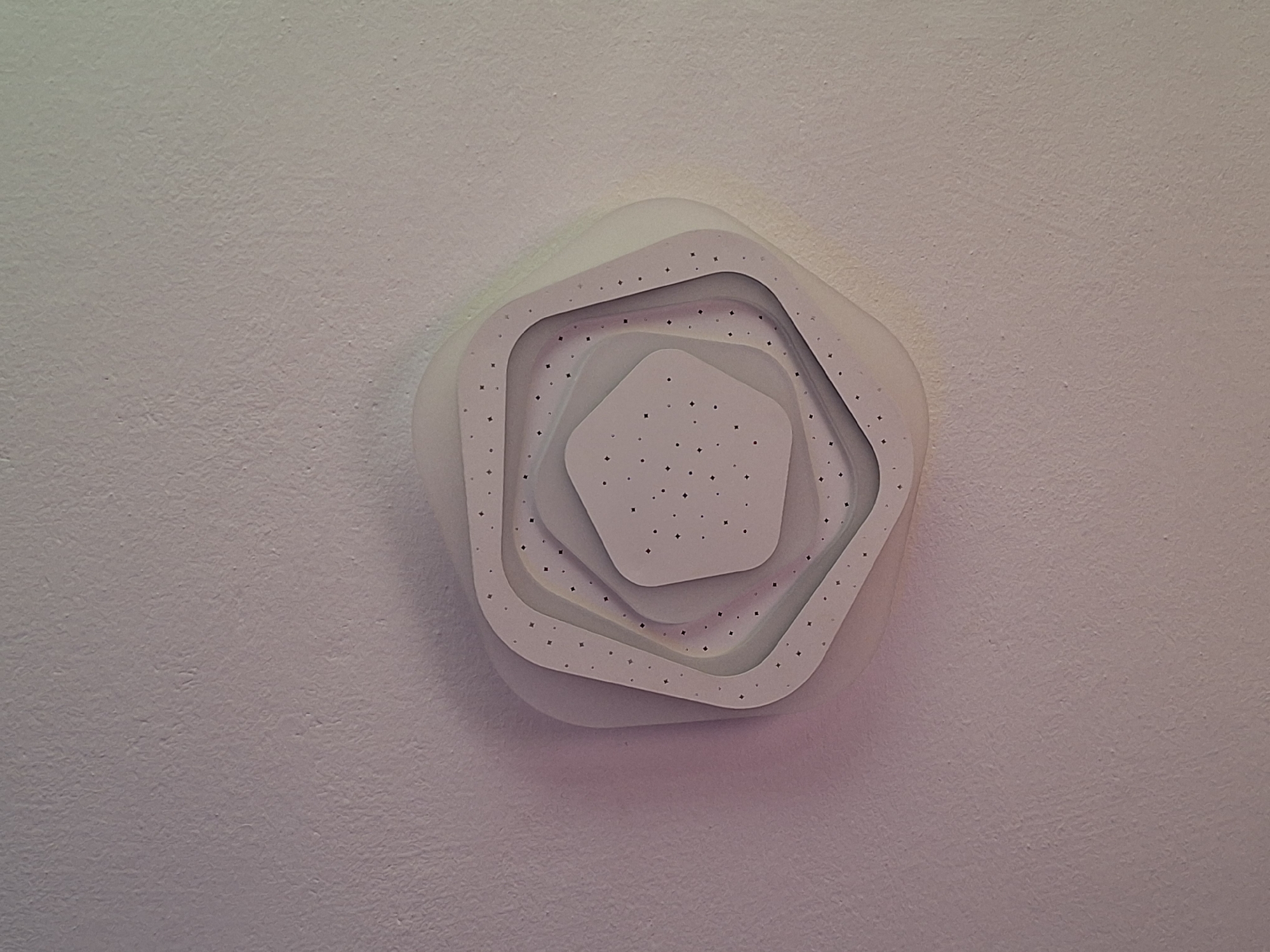 The image shows a ceiling fixture with a layered, hexagonal design, mounted on a textured, light-colored ceiling. The fixture has multiple concentric rings, each with a slightly different shade and a pattern of small, dark dots. The innermost layer is a solid, lighter hexagon. The lighting is soft and appears to be emanating from within the fixture, casting a gentle glow. The overall impression is of a modern, decorative light source. The location in the city of Varna, Bulgaria, is not visually discernible from the image.