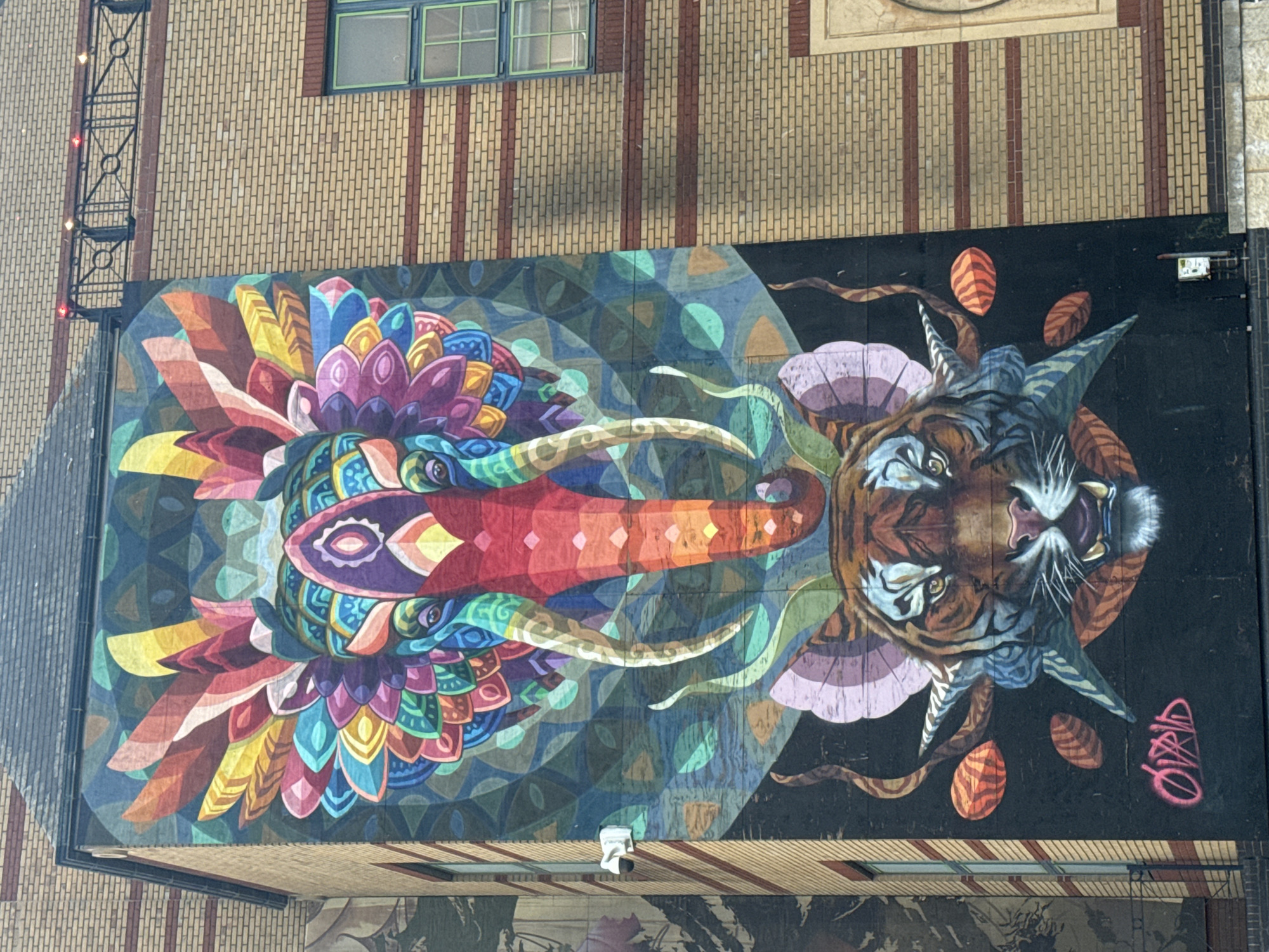 The image shows a large, colorful mural painted on the side of a brick building in Southend-on-Sea, United Kingdom. The mural depicts a stylized elephant's head above a tiger's head, both rendered in vibrant, abstract patterns and colors. The elephant's headdress is elaborate, with feather-like elements in shades of red, orange, yellow, blue, purple, and green. The elephant's trunk is orange and extends downwards. Below the elephant, the tiger's face is visible, with fierce eyes and bared teeth. The tiger also has abstract, wing-like elements behind its head in shades of purple and blue. The background of the mural is a geometric pattern in various shades of blue and green. The building itself is made of light brown bricks with darker red horizontal stripes. There are several windows visible on the building, one with green frames. A black metal railing with circular designs is present on an upper level. A white security camera is mounted on the wall to the left of the mural. A tag that appears to read "OPID" in pink graffiti is visible on the lower left portion of the mural. The overall scene is an urban outdoor environment.