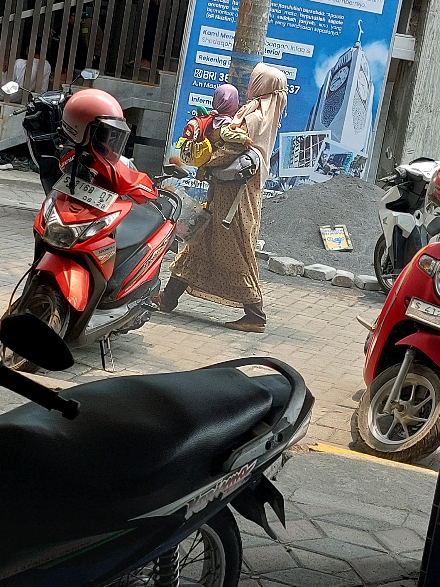 The image captures a street scene in Sumberrejo, Indonesia, under bright daylight. A woman wearing a hijab and a long, patterned dress is seen carrying a baby on her back. She is walking past a red motorcycle with a pink helmet on it, parked on a paved sidewalk. Next to the motorcycle, a pile of gravel and a white motorcycle are visible. A large blue poster with text and an image of a clock tower is displayed prominently in the background. The overall environment suggests an urban setting with pedestrian and vehicular traffic.