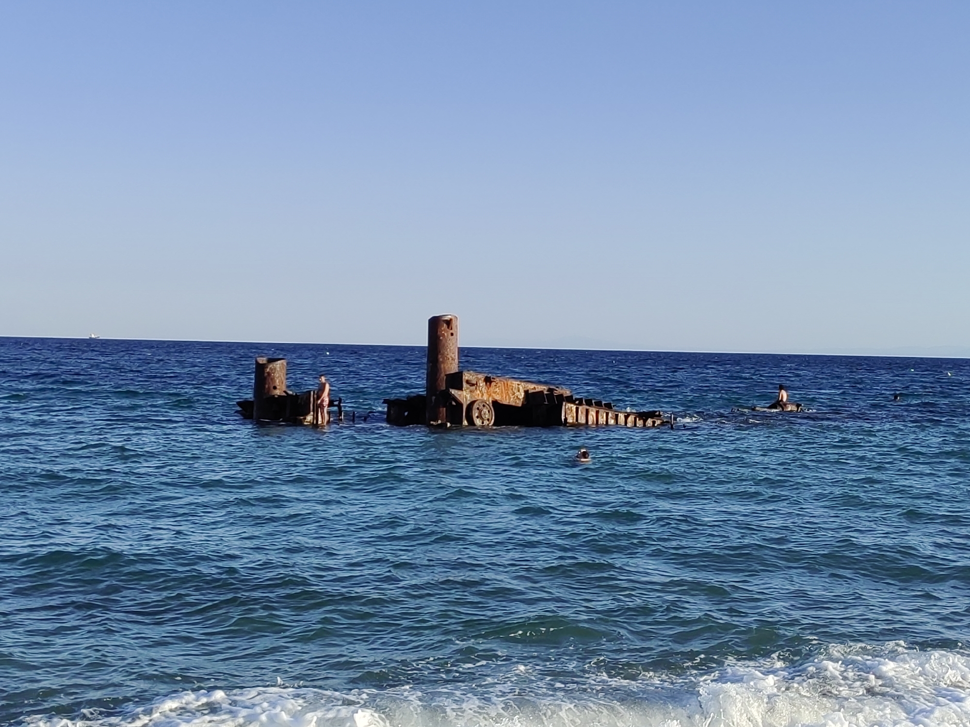 The image depicts the remnants of a shipwreck partially submerged in the sea off the coast of Perea, Greece. The wreckage, made of rusty metal, protrudes from the water, with some parts forming structures resembling pillars and platforms. A few individuals are visible in the water, some swimming near the wreck and others standing on parts of it. The sea is a vibrant blue with moderate waves and foam near the shore. The sky above is clear and blue, indicating a sunny day. In the distance, a small boat can be seen on the horizon. The scene conveys a sense of a historical site being used for recreational activities.