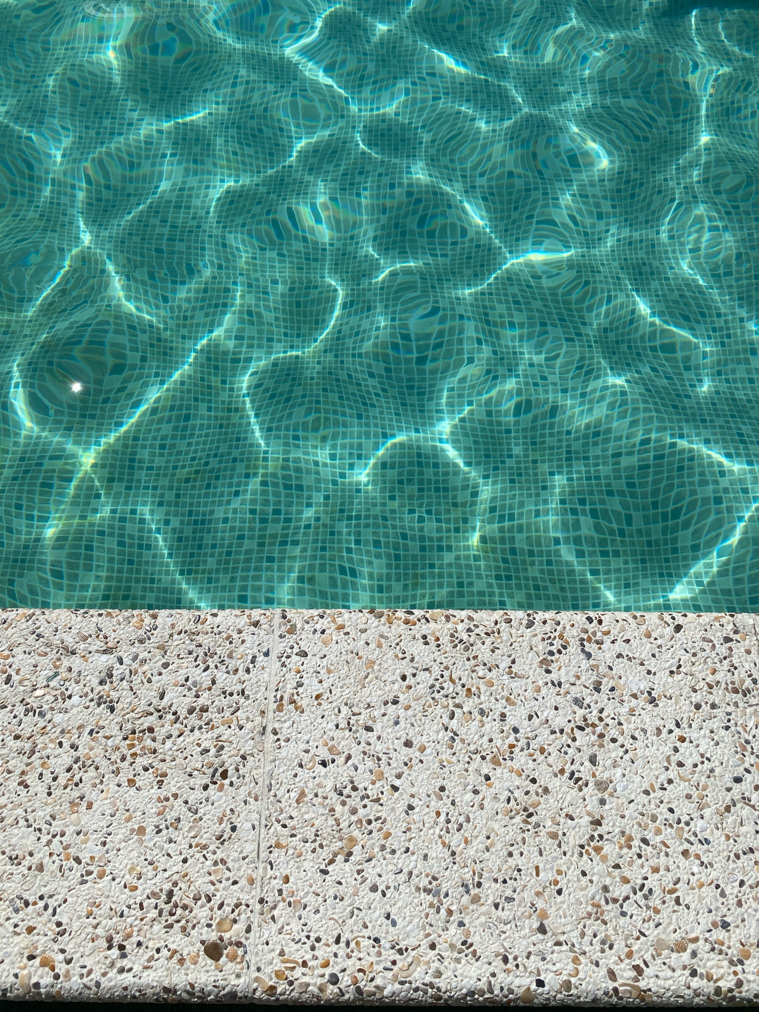The image is a close-up, top-down view of a swimming pool and its edge. The water is a clear turquoise color, with sunlight reflecting off the surface in shimmering patterns that distort the view of the underlying mosaic tiles. The mosaic tiles are small and square, forming a grid beneath the water. The pool edge is made of a textured material, appearing to be aggregate or exposed aggregate concrete, composed of small, multi-colored pebbles and shells embedded in a light-colored cementitious base. The edge forms a distinct horizontal line dividing the water from the surrounding deck. The scene is devoid of people, animals, or any discernible objects other than the pool and its immediate surroundings. The lighting suggests it is daytime, likely sunny. The location of this scene in the city of Nebug, Russia, is not visually indicated in the image itself.