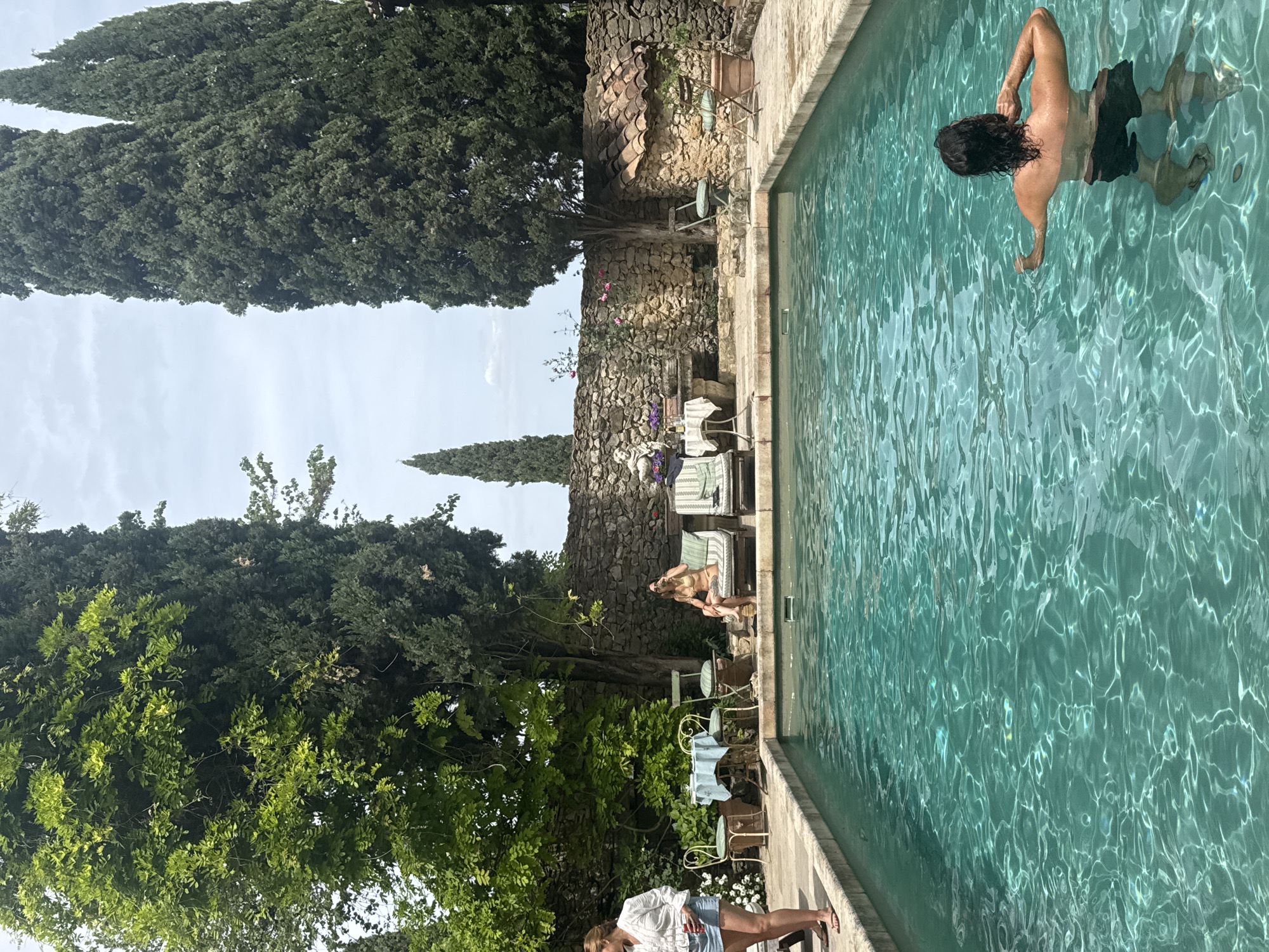 The image depicts an outdoor swimming pool with several people nearby. A man is swimming in the pool, facing away from the camera. A woman is sitting on a lounge chair next to the pool, and another woman is standing near the edge of the pool in the lower part of the image. The pool is surrounded by a stone wall, vegetation including tall, narrow trees and other plants and flowers. The setting appears to be in Saint-Victor-la-Coste, France, based on the style of the building and landscape. Chairs and tables are positioned along the edge of the pool, suggesting a relaxed or social setting.