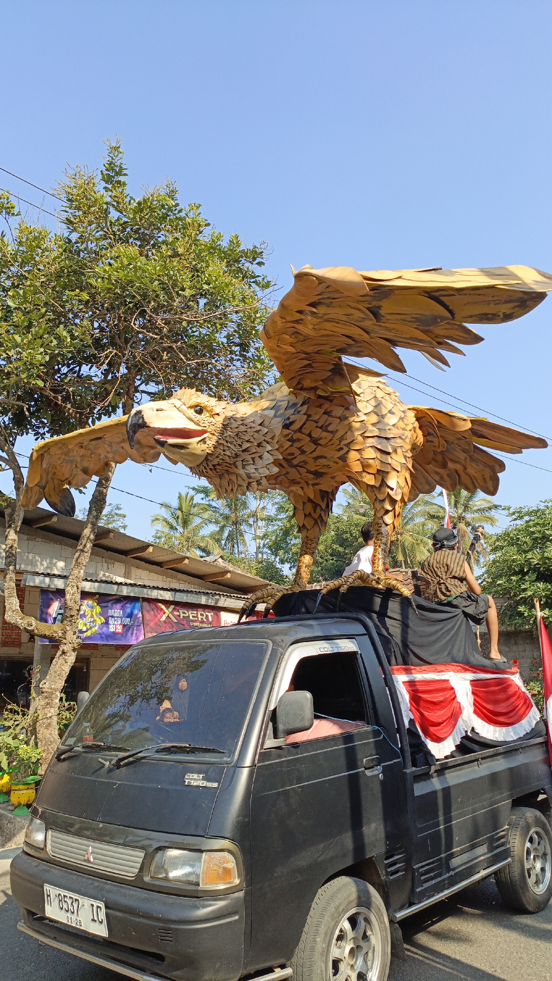 The image depicts a black pickup truck with a large, golden eagle sculpture mounted on its bed. The eagle sculpture is intricately detailed, with its wings spread wide and its beak open. Two individuals are visible on the truck bed, one seated and one standing. The truck is decorated with red and white fabric draping along the sides of the bed. In the background, there are trees, buildings, and a clear blue sky. The scene appears to be outdoors, possibly a street or a parade route. A license plate visible on the front of the truck reads "H-8537 IC" with a date "11-28". The location is identified as Krajan Lor, Indonesia.