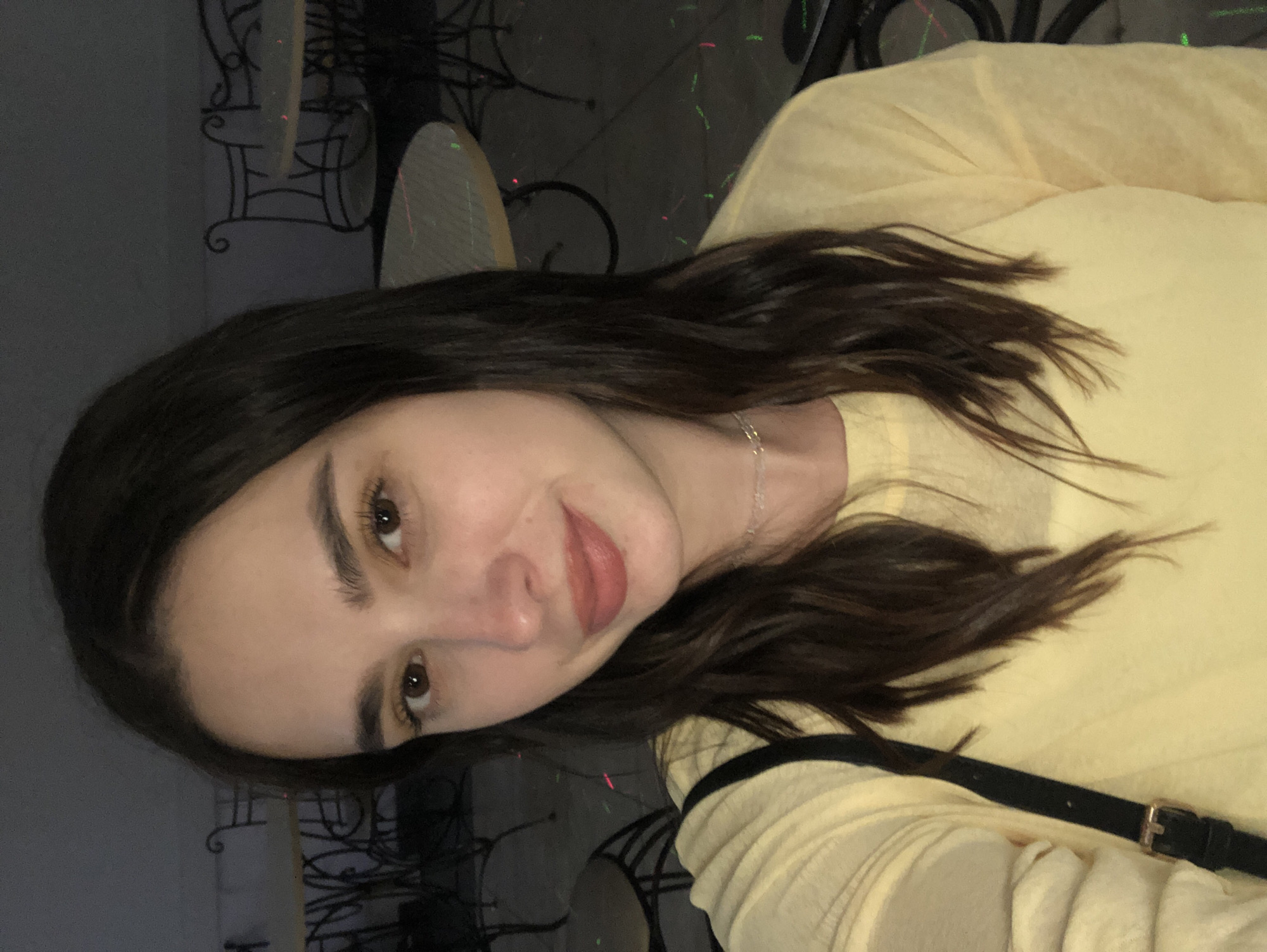 A young woman with dark brown hair and brown eyes is taking a selfie in what appears to be an outdoor seating area with tables and chairs. She is wearing a light yellow long-sleeved shirt and a delicate silver necklace. The background is dark, with some colorful lights reflecting on surfaces, suggesting it might be nighttime or a dimly lit venue. The woman has a slight smile. The location is not specified within the image itself, therefore it is not possible to provide information about it being in Izhevsk, Russia.