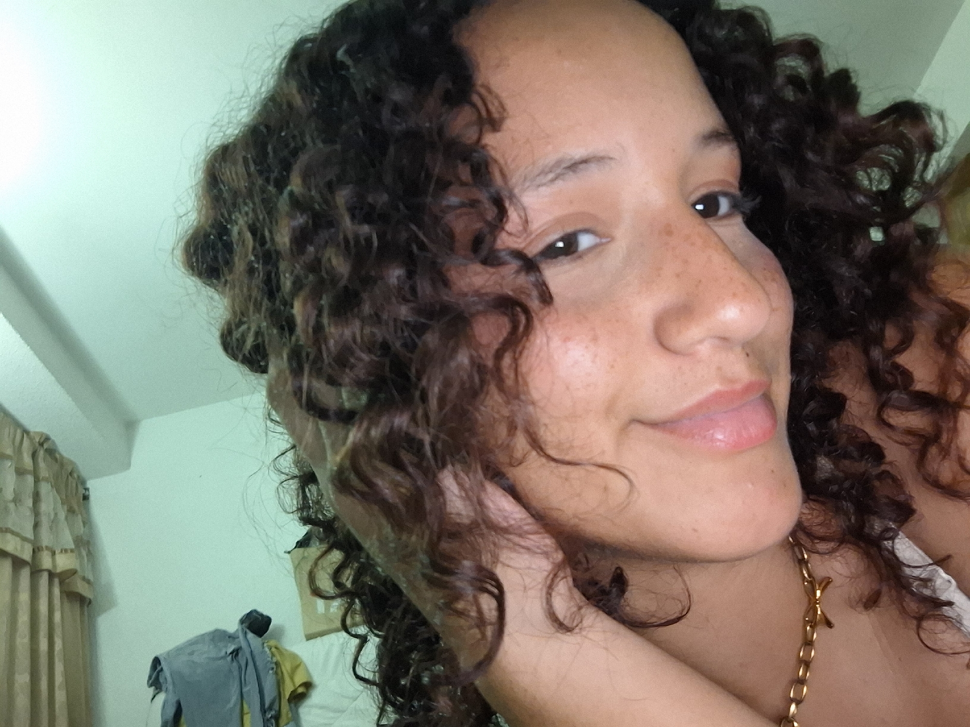 The image is a close-up, low-angle selfie of a woman with dark, curly hair and light brown skin. She is wearing a white top and a gold chain necklace with a "YSL" pendant. Her right hand is behind her head, and her left arm is resting on her chest. Her eyes are brown, and she has light freckles on her face. She is smiling slightly, with her lips slightly parted. The background appears to be a bedroom with a light blue wall, a beige curtain, and some clothing piled on a surface. The image is likely taken in Juan Griego, Venezuela, given the context provided.