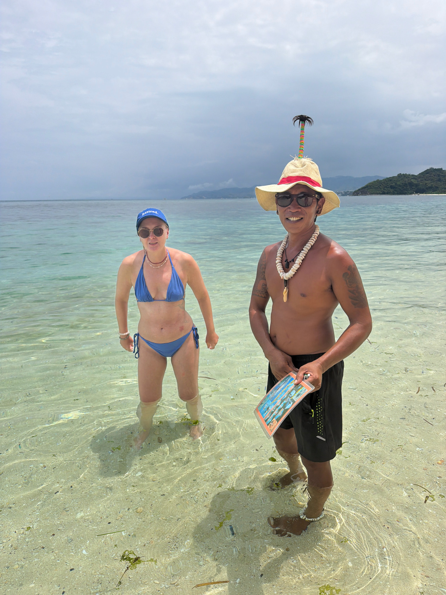 Two individuals are standing in shallow, clear turquoise water near a sandy seabed. On the left, a person wearing a blue bikini, sunglasses, and a blue baseball cap with the word "retired." visible, is looking towards the camera. On the right, a person wearing a straw hat adorned with a small, colorful palm tree, sunglasses, and a white shell necklace stands shirtless in black shorts. This individual is also looking at the camera and holding a rectangular object, possibly a brochure or map, with colorful images. The background shows a calm ocean with a distant, tree-covered island under a cloudy sky. This scene is likely on one of the beaches of Boracay Island in Malay, Philippines, known for its clear waters and white sand.
