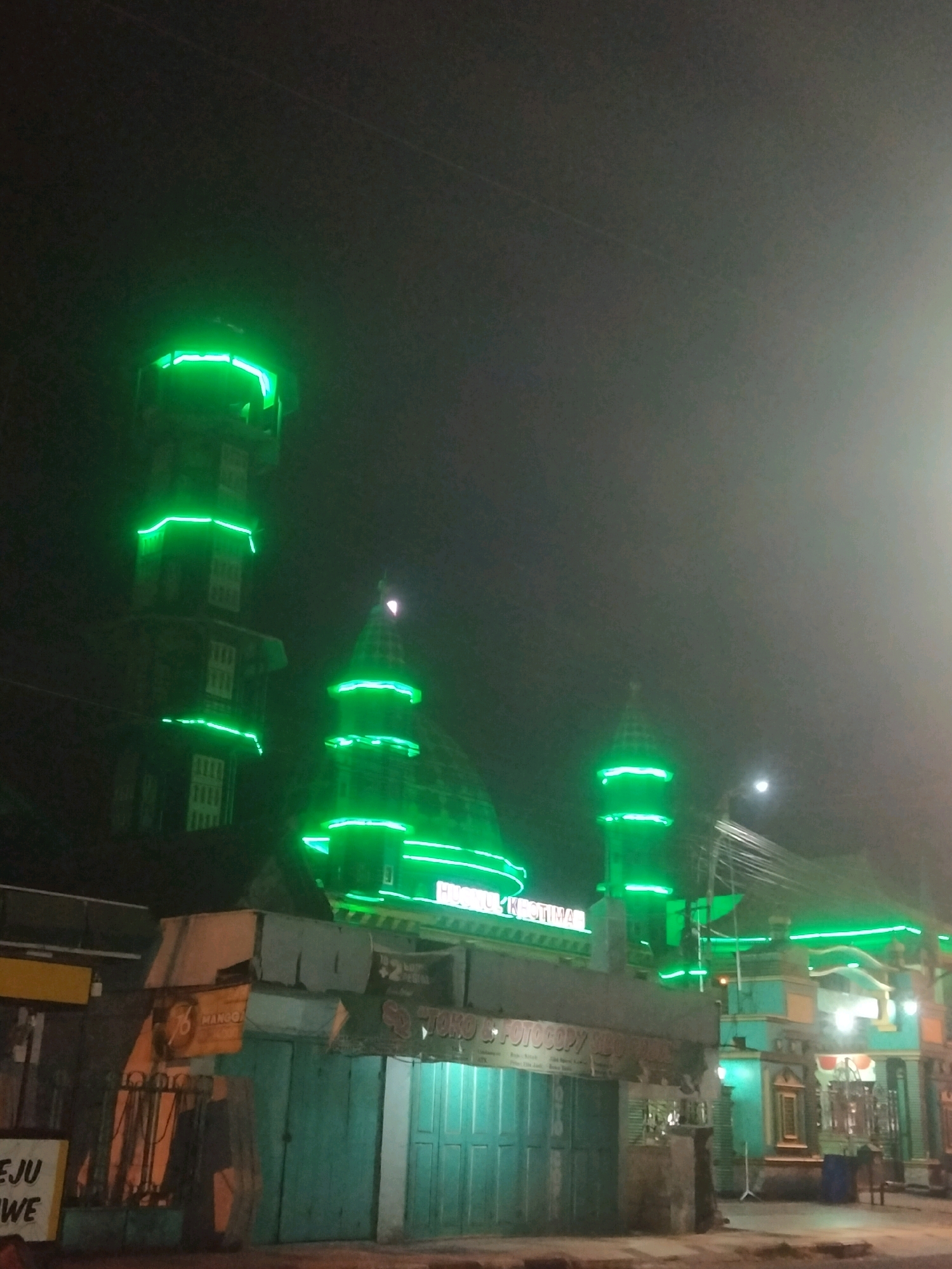 The image depicts a night scene in Merakurak, Indonesia, featuring a mosque or religious building illuminated with bright green neon lights. The building has multiple domed structures and minarets, all outlined with the vibrant green lighting. In the foreground, there are closed storefronts and signs, suggesting a commercial area. The sky is dark, with a faint moon visible. The overall impression is one of a nocturnal urban landscape dominated by the striking illumination of the religious structure.