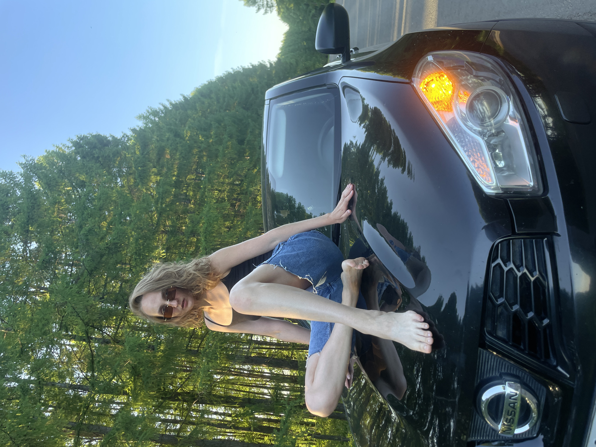 A woman wearing a black top, denim shorts, and sunglasses is sitting on the hood of a black Nissan SUV. The car is parked on a road alongside a forest. The car's right turn signal is illuminated. The location is likely Подшивалово, Russia, given the user's prompt.