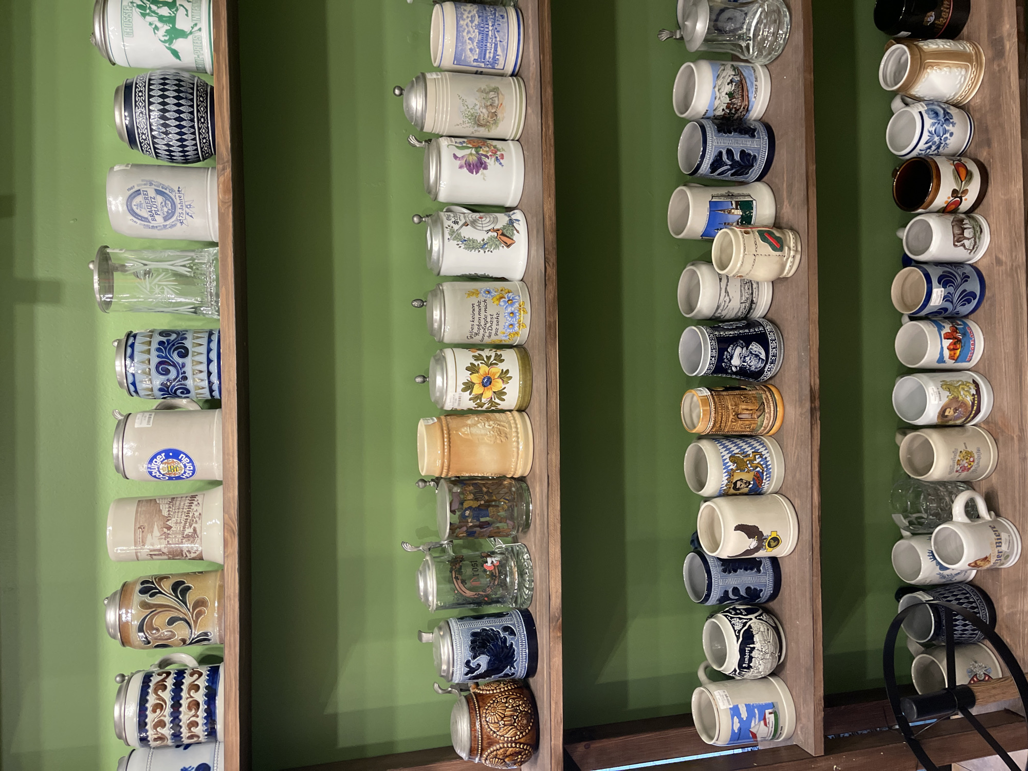 The image shows a collection of ceramic and glass steins displayed on wooden shelves against a green wall. The steins are of various sizes, colors, and designs, featuring scenes, patterns, and text. They are arranged in rows on two shelves. The top shelf holds eight steins and a glass mug. The bottom shelf displays fifteen steins and a glass mug. The lighting suggests an indoor setting, possibly a shop or a private collection. The image does not contain any people or any recognizable landmarks that would indicate a specific location in Svetlogorsk, Russia.