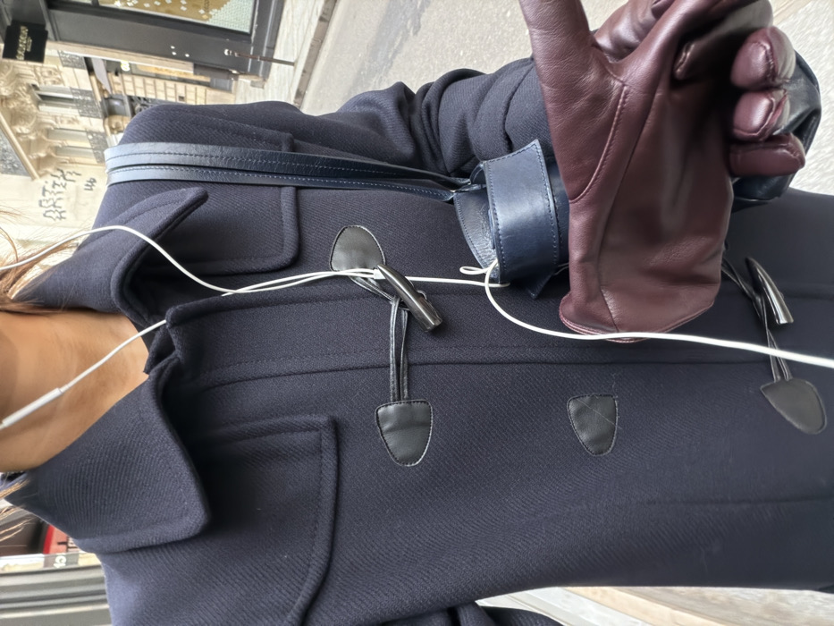 The image presents a close-up view of an individual from the neck to the waist, positioned on an urban street in Paris, France. The person wears a dark navy blue duffle coat, fastened partially with two visible black leather patches and metallic toggles. White wired earphones are draped around the neck, with the wire extending down the front of the coat. A dark navy leather shoulder bag, identifiable by its strap, is carried on the person's right side. The right hand, clad in a burgundy leather glove, is held in the foreground.
The background features a light-colored Haussmannian-style building with ornate architectural details, including a partial view of a balcony railing on an upper floor. A ground-level storefront with dark glass and faint white lettering, possibly indicating "SAINT LAURENT," is visible. Graffiti, including the word "ARTIZ," is present on the stone facade above the storefront. The street or sidewalk in the lower right background is paved in a gray tone, consistent with an urban setting.
