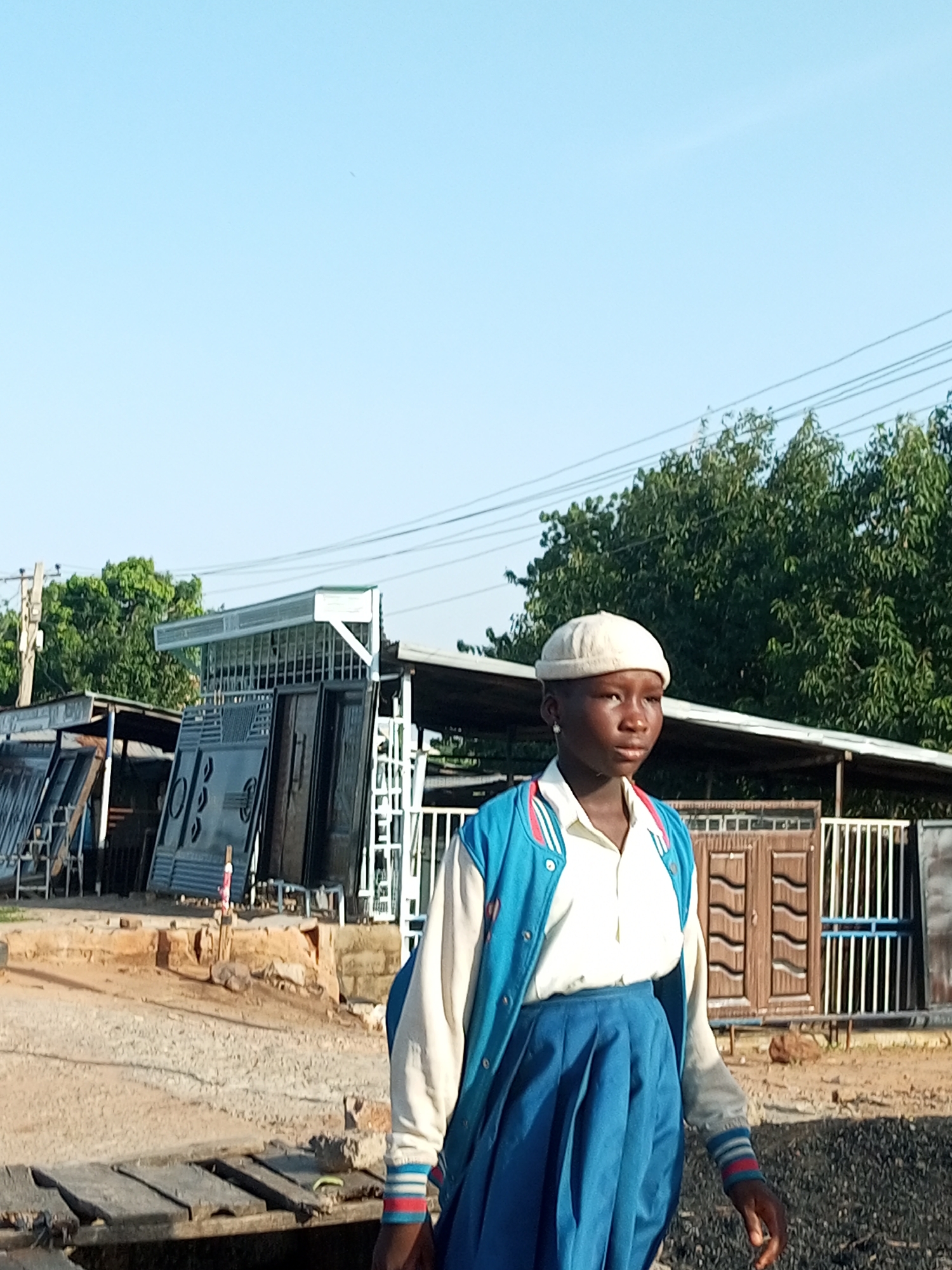 A young African female, wearing a white skullcap, a white collared shirt, a blue and white varsity jacket with red and white striped cuffs, and a blue pleated skirt, stands facing right in the foreground. She is in a market setting in Garko, Nigeria, with a clear blue sky overhead. Behind her, a stall displays various metal gates and doors. In the mid-ground, there are more stalls with metal products, trees, and utility poles with electrical wires. The ground is a mixture of dirt, gravel, and some wooden planks. The overall impression is an outdoor market scene during daylight.