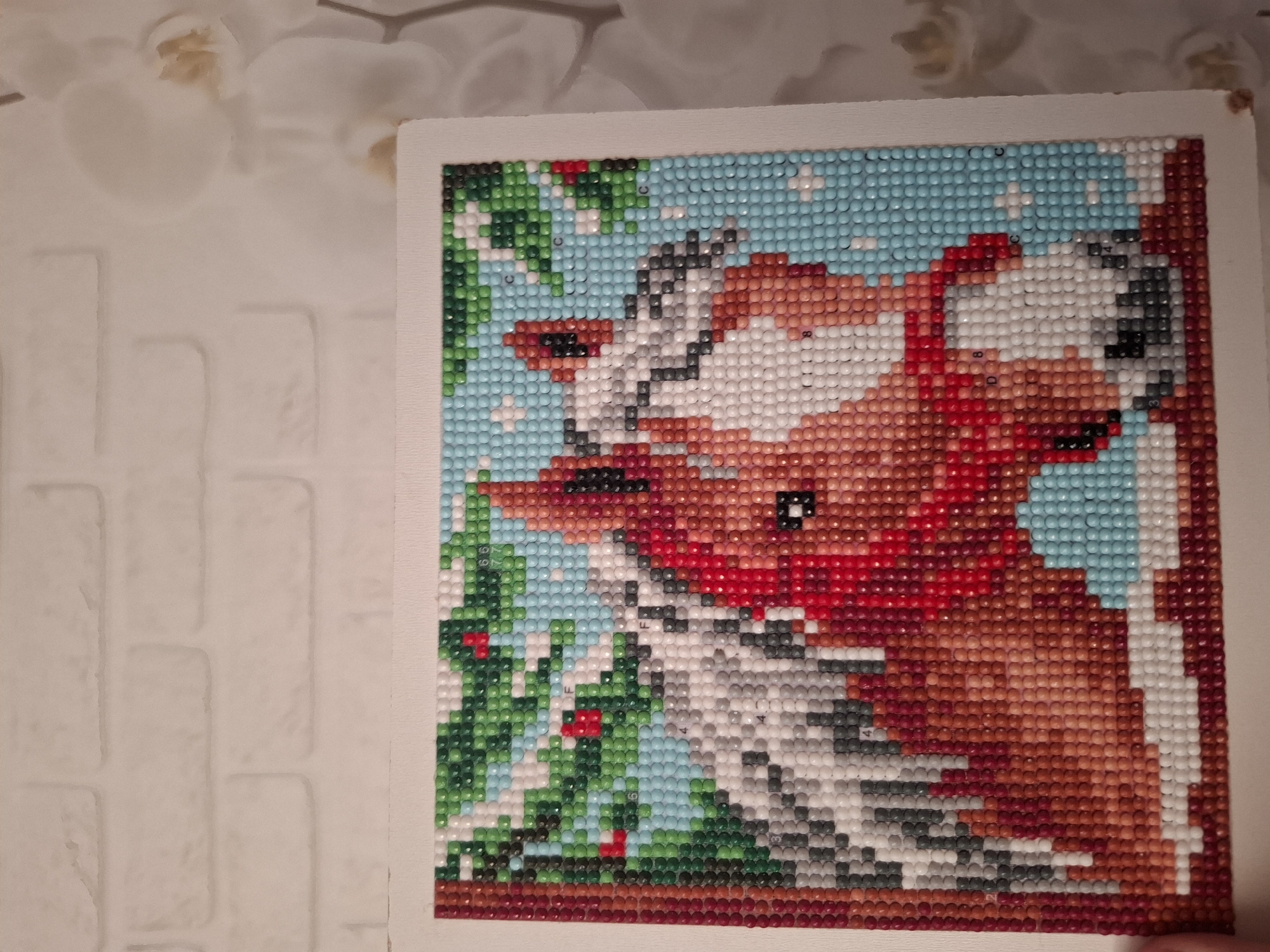 A square diamond-painting image depicting a reddish-brown and white fox with a red scarf, with holly branches on the left side of the frame, is positioned on a white surface. The image is close-up. The background of the image is a light blue diamond-dot pattern. The location is unknown.