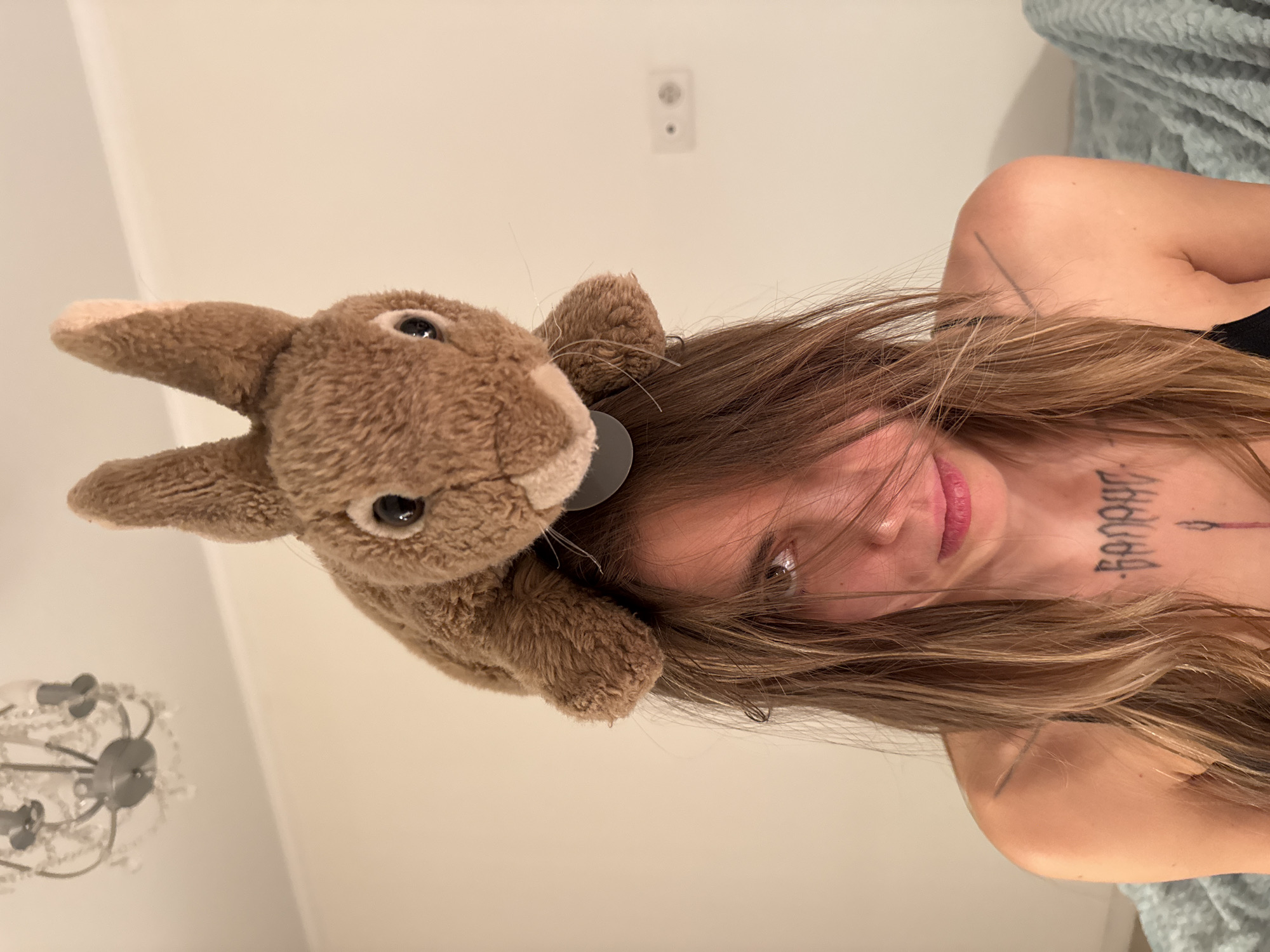 In an indoor setting, a young woman with long, light brown hair is positioned centrally, looking towards the camera with a somber expression. Her hair partially obscures her face. A brown stuffed rabbit is placed on top of her head. She is wearing a black top, revealing tattoos on her chest and collarbone. The background is a plain, light-colored wall. A chandelier is visible in the upper left corner, and a bed with a textured blue blanket is to the right. The presence of an electrical outlet on the wall suggests a residential interior. There is no indication of the location being Moscow, Russia.