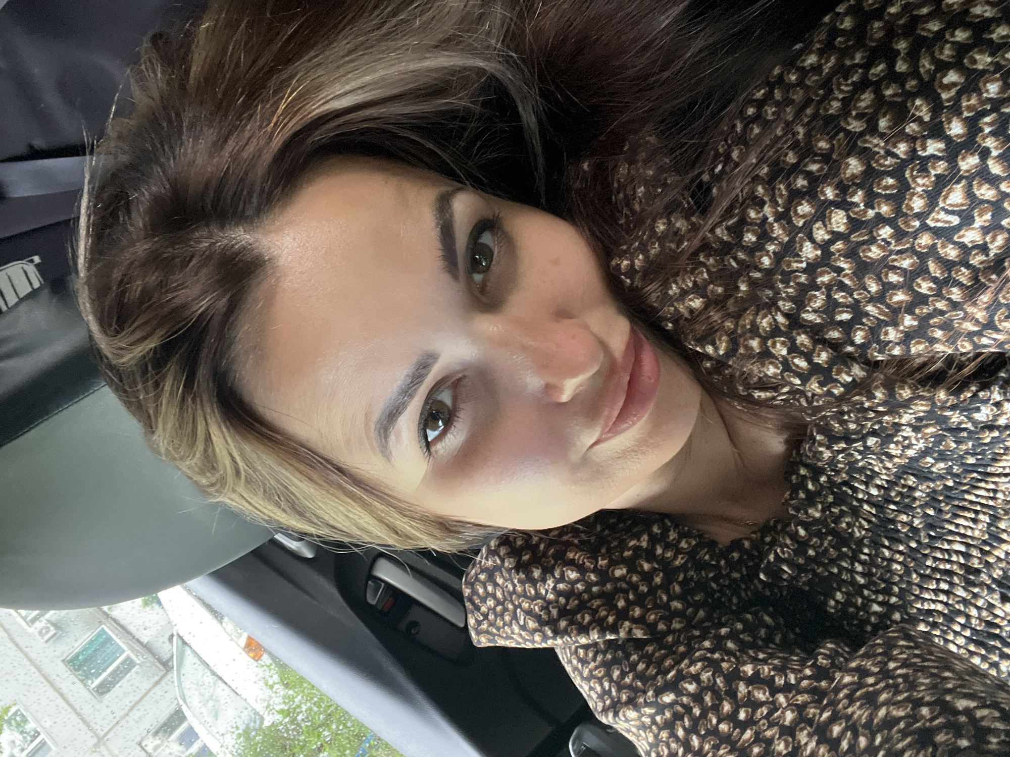 A woman is taking a selfie in a car. She has long brown hair with blonde highlights and is wearing a brown and black leopard print top. The background shows a slightly blurred view of a building with windows and some trees outside, suggesting an urban environment. The image appears to be taken during the day, possibly with overcast skies, as indicated by the lighting.