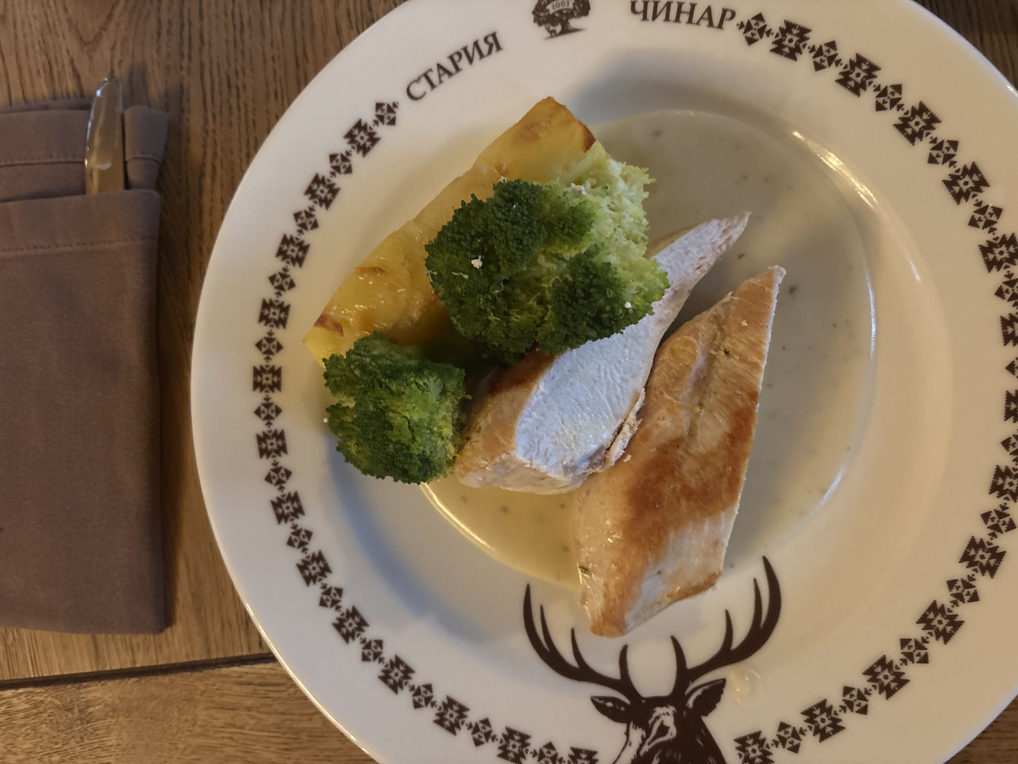 The image depicts a plate of food, likely chicken breast with broccoli and potatoes, served in a creamy sauce. The plate has a decorative pattern around the rim and a deer's head motif at the bottom. The plate is on a wooden surface. To the left, there is a brown cloth napkin with cutlery inside. The plate's rim is imprinted with text that reads "СТАРИЯ ЧИНАР". The restaurant is located in Sofia, Bulgaria.