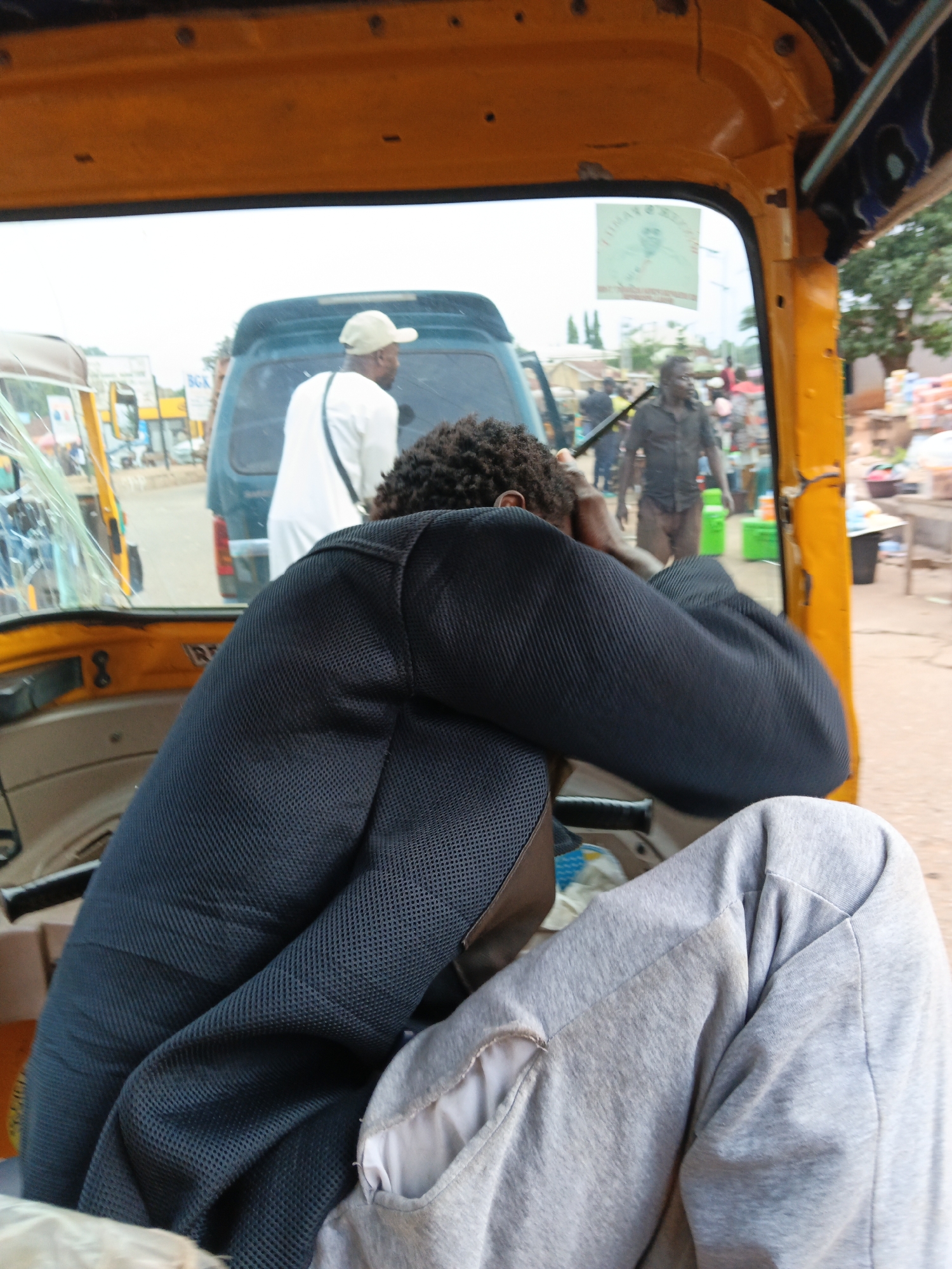 The image shows the interior of a yellow auto-rickshaw, looking out through the rear window. In the foreground, the back of a person wearing a dark, mesh-like long-sleeved shirt and light grey pants is visible, with their arms crossed over their chest and head bowed. Behind them, through the window, a man wearing a white shirt and a beige cap is seen from the back, walking away. Further in the background, a man in a dark shirt and light brown pants is walking. The environment appears to be an outdoor urban setting, possibly a street or market, with buildings and stalls visible. The overall lighting suggests daylight.