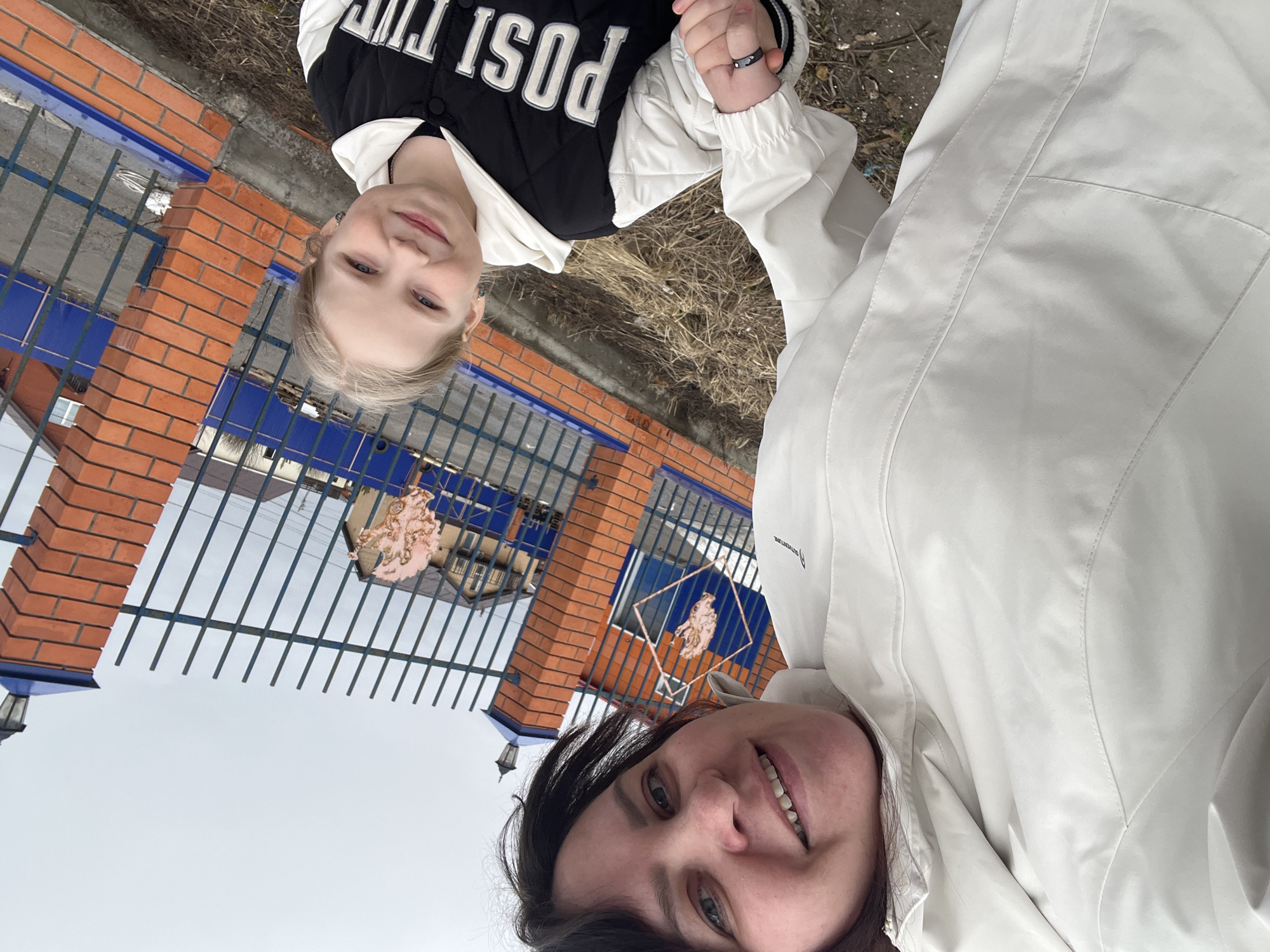 The image, oriented sideways, shows an adult and a child outdoors in Protasy, Russia, smiling at the camera. The adult, positioned in the foreground to the right, has dark hair and a warm smile. She is wearing a light-colored, possibly white, jacket. Text visible on her jacket in a cropped view reads "ADVENTURE." Her hand is extended towards the child's.
The child, a girl with blonde hair, stands to the left of the adult and also smiles. She wears a black and white bomber-style jacket with the partially visible text "POST T..." on the front. She has a small earring in her ear and appears to be holding hands with the adult.
The background features a fence constructed from reddish-brown brick pillars and dark metal bars. Behind the fence, blue structures or panels are visible, and an ornate, golden-toned emblem depicting a horse-like figure within a diamond shape is attached to the fence. The ground is covered with dry grass and patches of dirt, suggesting a natural, somewhat unkempt outdoor area. The sky overhead is overcast, indicating a cloudy daytime. Both individuals appear happy and comfortable.
