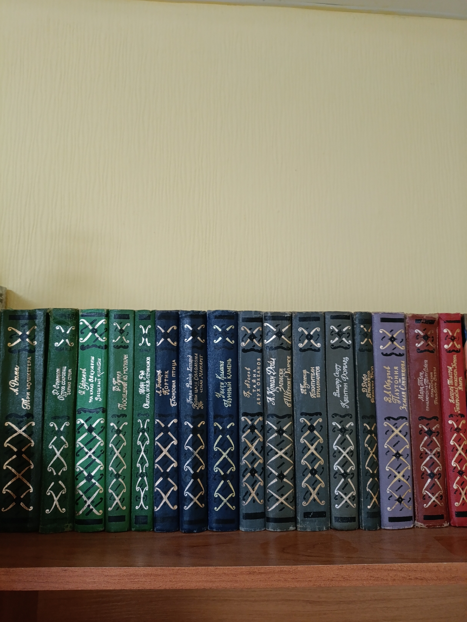 A straight-on shot depicts a bookshelf filled with at least fifteen books with decorative spines, photographed in Chaikovsky, Russia. The books are arranged closely together on a brown wooden shelf. The spines are predominantly green, blue, purple, and red, each adorned with a repeating "X" or crossed pattern in a lighter color. Each book has Russian text printed on the spine. The background is a plain, light yellow wall with a subtle textured pattern. The books sit near the bottom of the frame, leaving a large expanse of the wall visible.