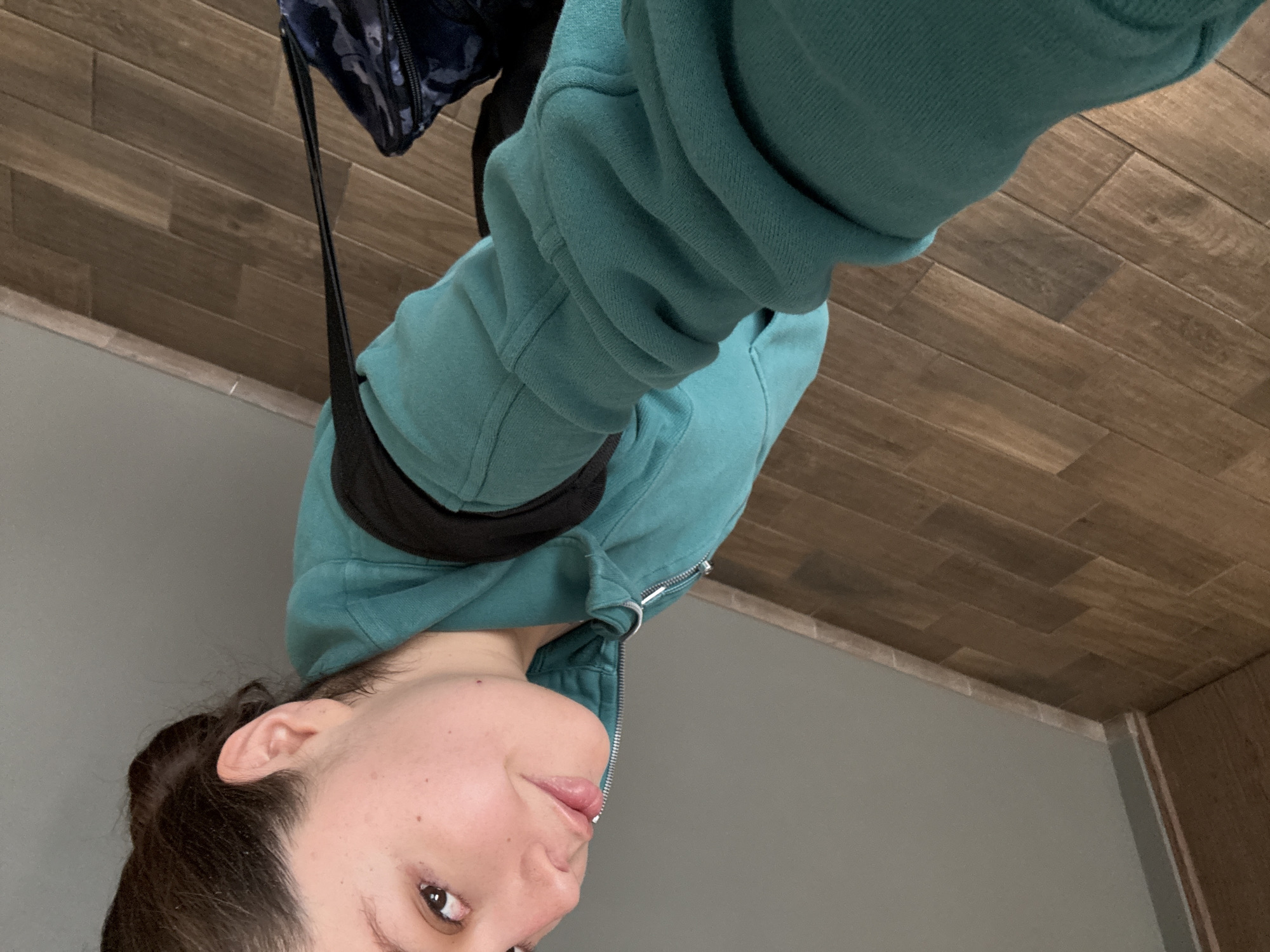 A person, appearing to be a young adult female, takes a selfie indoors. The image is oriented with the person's head facing downwards and to the left, but if rotated correctly, she would be looking slightly upwards at the camera. She is wearing a teal or turquoise hooded sweatshirt, which is partially unzipped, revealing a silver zipper. A black strap, likely from a bag, crosses her upper body. A dark blue or black patterned bag is visible in the upper right corner of the frame (when rotated correctly), hanging from this strap.
The visible portion of her face, primarily the right side, shows her looking directly towards the camera with a neutral to slightly pleasant expression. Her dark hair is pulled back, and her right ear is visible.
The background features multiple elements. What appears to be the ceiling is covered with wood-patterned tiles or planks in varying shades of brown. To the left of the person is a plain light grey wall, and to the right, a vertical wooden panel or door frame is visible. The setting appears to be an indoor space with modern finishes. There is no visible text in the image. The location is provided as Mexico City, Mexico, although this detail is not visually discernible within the photograph.