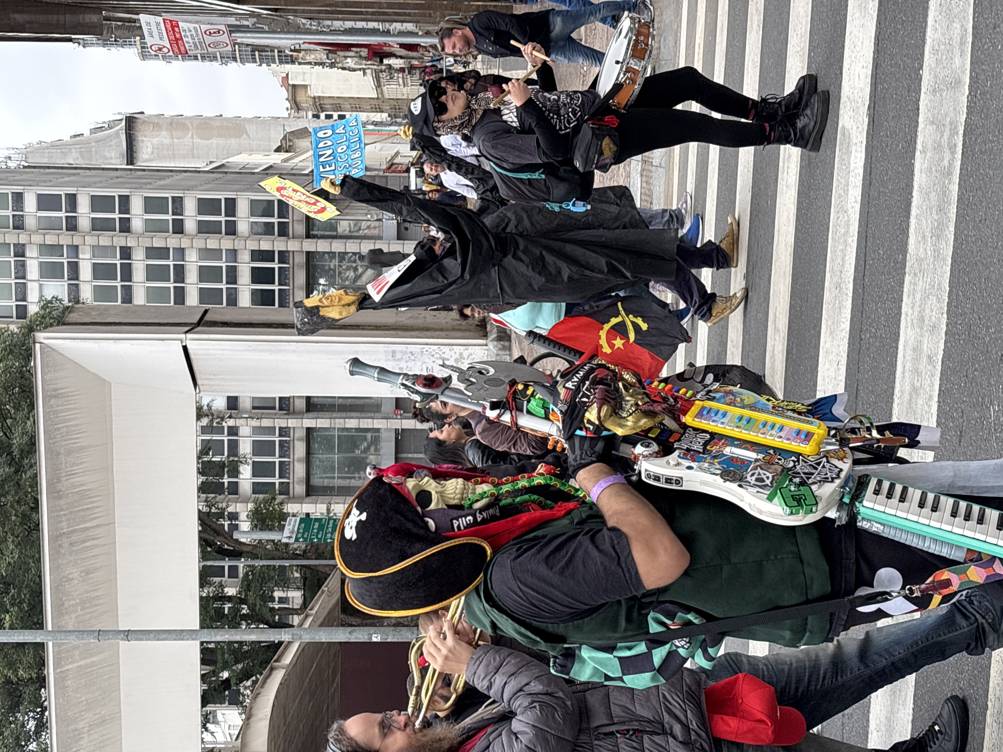The image depicts a street protest in what appears to be the city of São Paulo, Brazil. A group of people is marching on a zebra crossing in the middle of a road. One person in the foreground is dressed as a pirate, wearing a black hat with a skull and crossbones, playing a brass instrument (possibly a trumpet). The pirate has a keyboard, decorated guitar, and a skull mask. Behind the pirate, other protesters are visible. One wears a black cloak and mask, holding up two signs, one reading "Vendo Escola Publica", the other with the words "Enzo" and "Roulag wild". Another protester plays a small drum. A building facade is visible in the background.