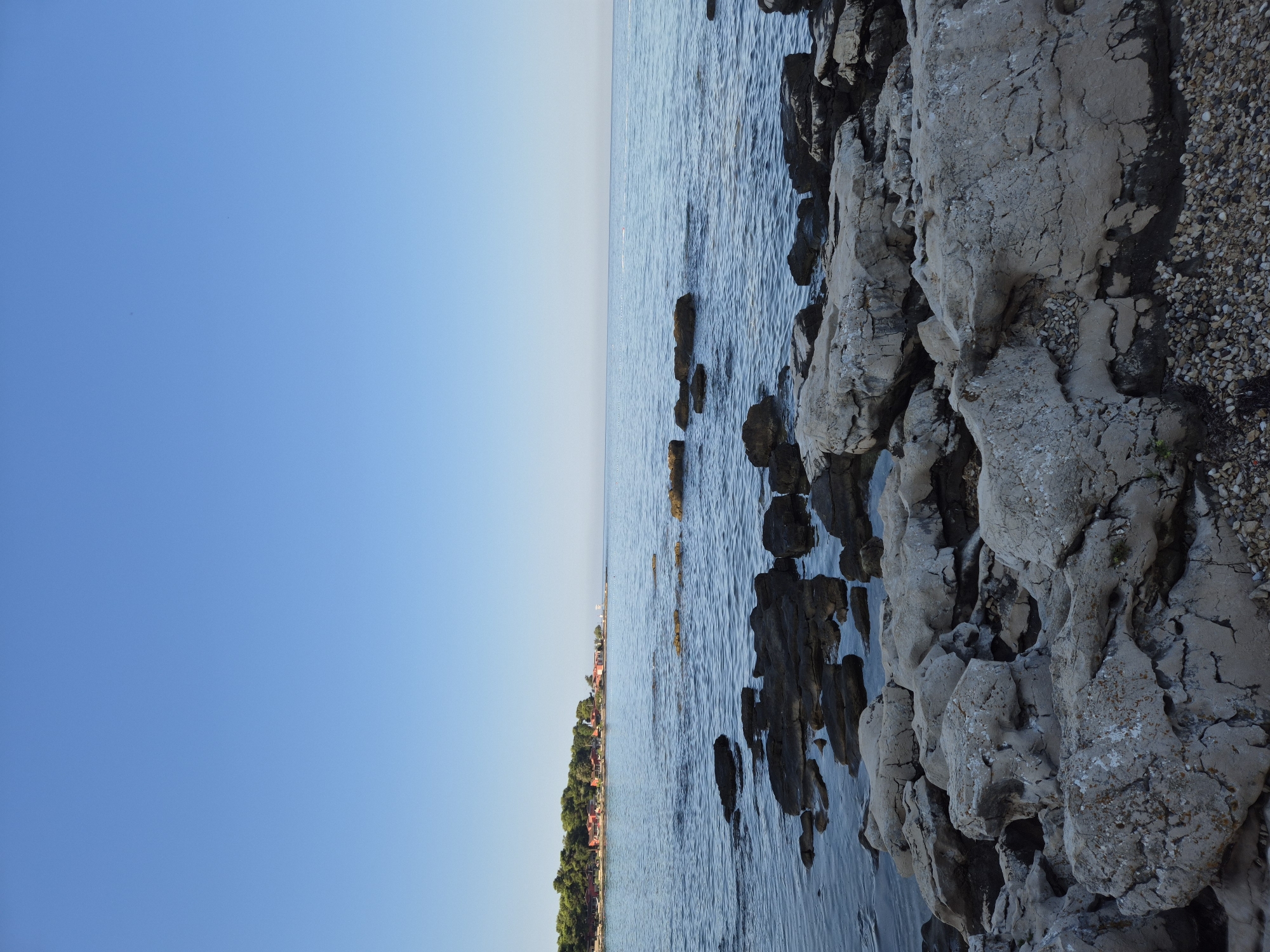 An outdoor, vertical, eye-level shot captures a rocky shoreline meeting a calm, blue sea under a clear, light blue sky. The foreground is dominated by large, grey, rough-textured rocks scattered across a pebbly beach. The middle ground features darker, submerged rocks and ripples on the water's surface. In the background, a distant treeline with scattered red-roofed buildings and a thin line of green vegetation marks the coastline. The sea extends to the horizon where it meets the sky. This image depicts the coast of Monterol, Croatia. There are no people visible in the scene.