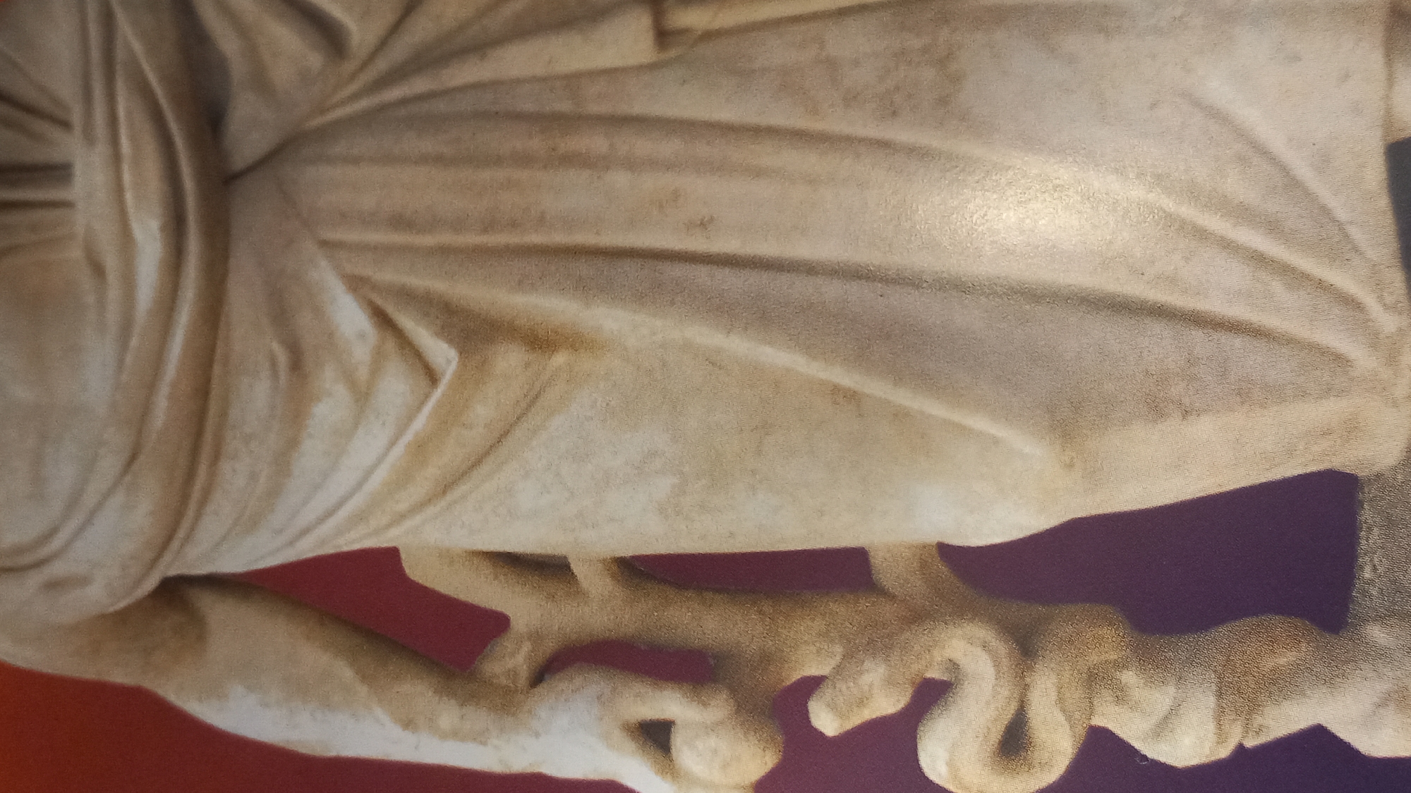 The image displays a close-up of a marble statue, focusing on its lower torso and arm, which holds a caduceus. The statue is draped in flowing robes, rendered with detailed folds and textures. The caduceus, a staff entwined by two snakes, is a prominent object held by the figure. The background transitions from a reddish hue at the top to a purple hue at the bottom. This statue is part of the House of the Masks in the city of Kos, Greece, a significant archaeological site.