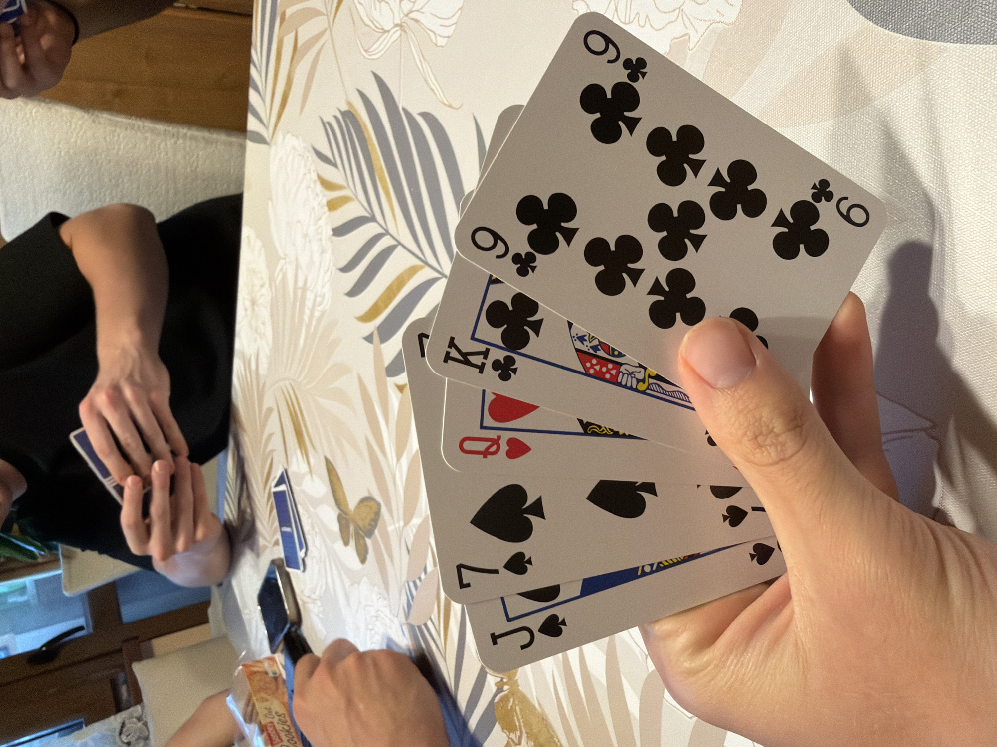 A person holds five playing cards in their right hand. The visible cards are a 9 of clubs, another 9 of clubs, the King of clubs, the Queen of hearts, a 7 of spades, and a Jack of spades. Another person sits to the left with a deck of cards in their hands. There is a pack of cookies visible on the table. The scene is indoors, on a patterned tablecloth. There is no evidence that the image was taken in Burgas, Bulgaria.